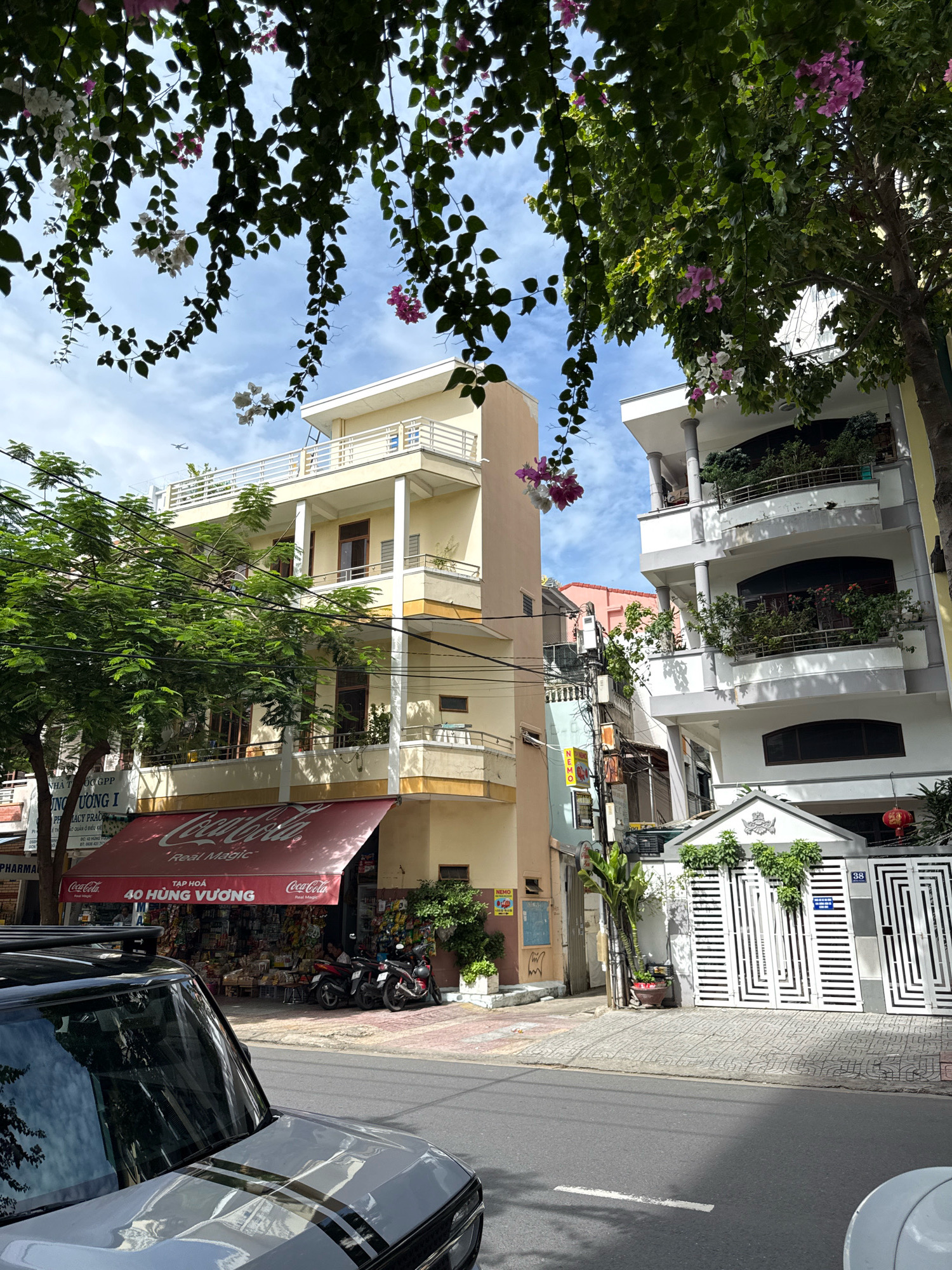 This is a street-level, eye-level, medium shot captured in Nha Trang, Vietnam, on a sunny day with a few clouds in the sky. The foreground is dominated by the hood and windshield of a gray SUV, reflecting the surrounding buildings and trees. Behind the SUV, a street runs parallel to the buildings. On the right side of the street, a white gated fence with a decorative top encloses a property. A red lantern hangs from the right side of the building above the gate. To the left of the gate, a multi-story building with a pale pink facade is visible. Further left, a shop with a red awning displaying the Coca-Cola logo and the text "40 HÙNG VƯƠNG" is situated. Behind the shop, a variety of goods are displayed on shelves. Two motorcycles are parked in front of the shop. Directly behind and to the left of the shop, a tall, slender, cream-colored building with balconies on multiple levels rises. To the far right of the frame, another multi-story building with a white facade and balconies filled with plants is prominent. Lush green trees and flowering vines with pink and white blossoms hang from above, partially obscuring the sky and the upper floors of the buildings. An airplane is visible in the sky above the cream-colored building. The overall scene depicts a typical urban streetscape in Nha Trang, with a mix of residential and commercial buildings, vegetation, and vehicles.