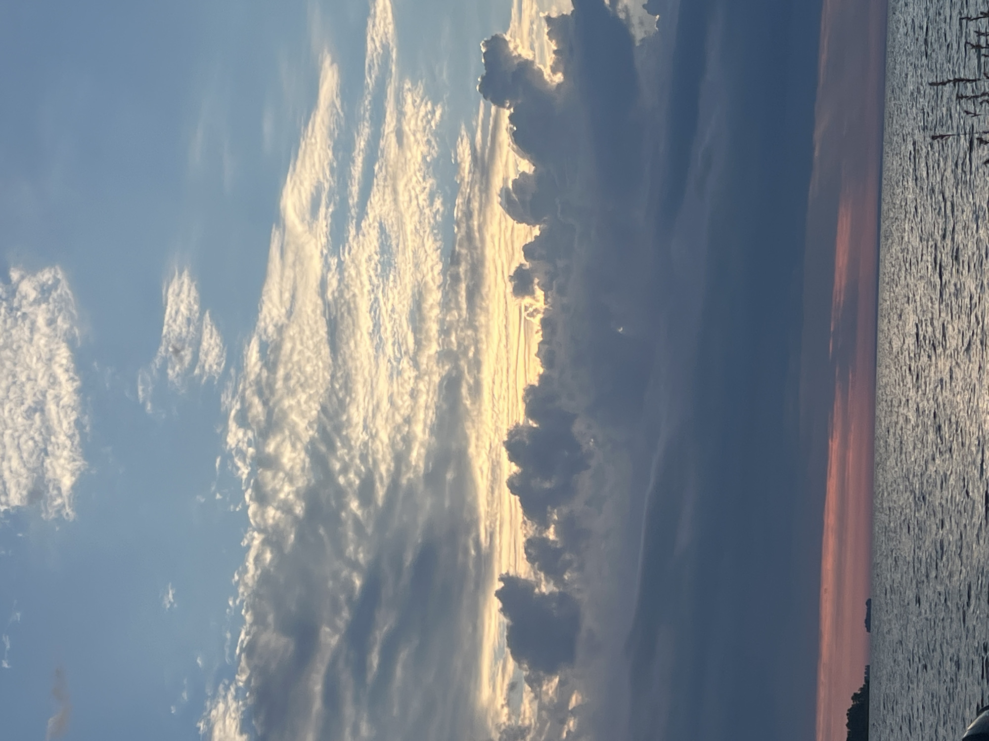 The image captures a serene seascape at dusk. The sky is filled with a dramatic display of clouds, ranging from light wispy formations near the top to darker, more solid masses closer to the horizon. A band of warm, golden light emanates from behind the clouds, suggesting the setting sun. Below the clouds, a vast expanse of water stretches across the lower half of the frame, reflecting the muted colors of the sky. A faint pinkish hue can be seen along the horizon, where the sky meets the water. On the left side of the image, a dark, silhouetted landmass with trees is visible, hinting at a distant shore. In the water, a small, dark object, possibly a boat or a structure, is positioned near the center. The overall impression is one of peaceful natural beauty.