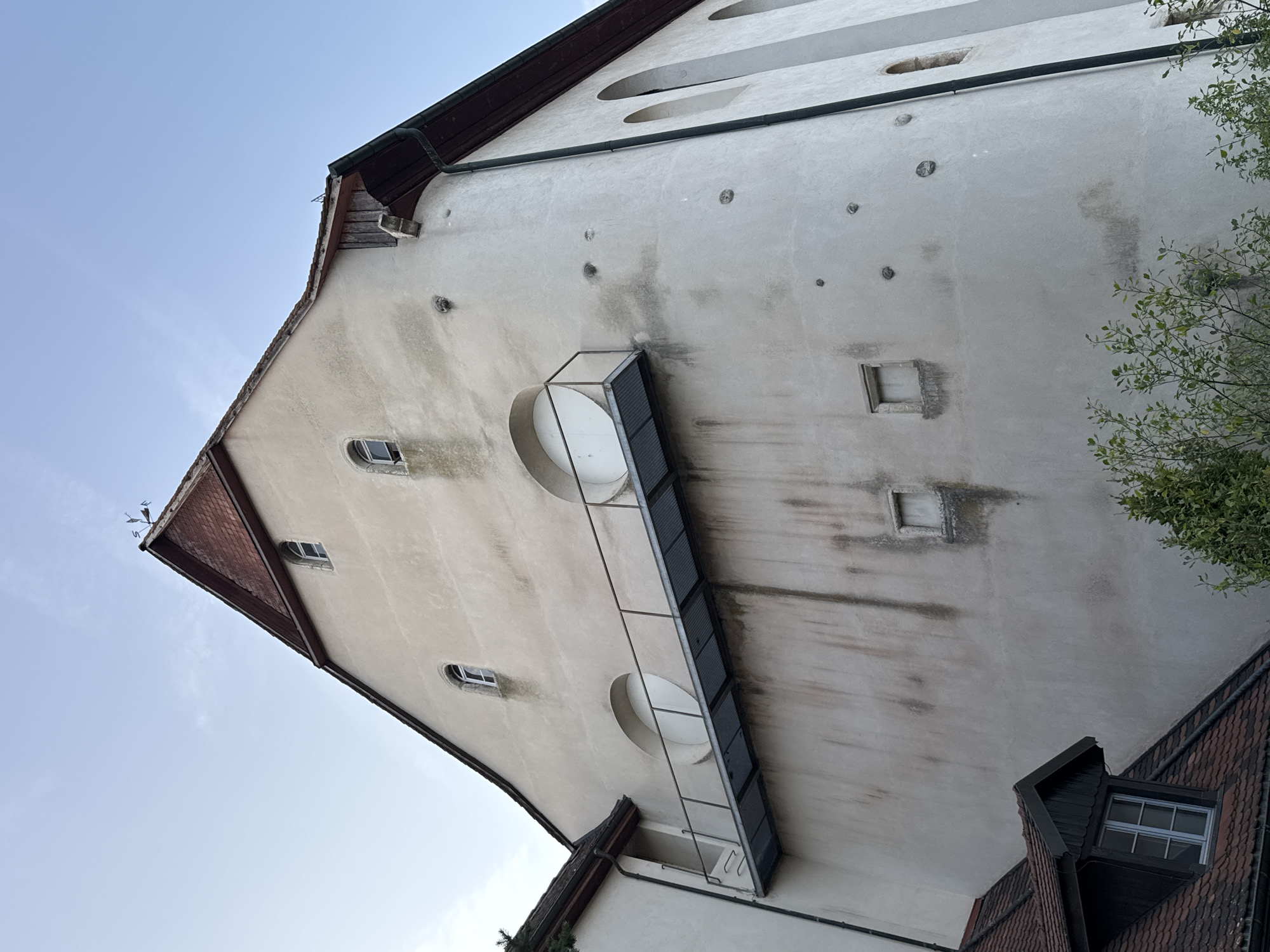 The image captures a portion of an old, light-colored building, possibly a former industrial or public structure, located in Porrentruy, Switzerland. The building has a steep, red-tiled roof with a visible weather vane at its apex. The facade features several architectural elements, including arched windows on the right side and a prominent metal walkway or balcony extending horizontally across the middle of the building. This walkway has a grated surface and a simple metal railing.
On the main section of the wall, there are two large, circular openings, each covered by a white panel. Smaller, square-shaped windows are also visible at different levels on the facade. The wall itself shows signs of age and weathering, with dark streaks and discoloration, possibly from water runoff or historical damage. Several small, circular indentations or fixtures are scattered across the wall's surface.
In the foreground, a section of another building with a dark-tiled roof and a dormer window with a multi-paned glass is visible. Green foliage from trees or bushes partially obscures the lower portion of the main building. The sky above is a clear, light blue, suggesting daylight. The overall impression is of an aged, historical structure in an urban setting.