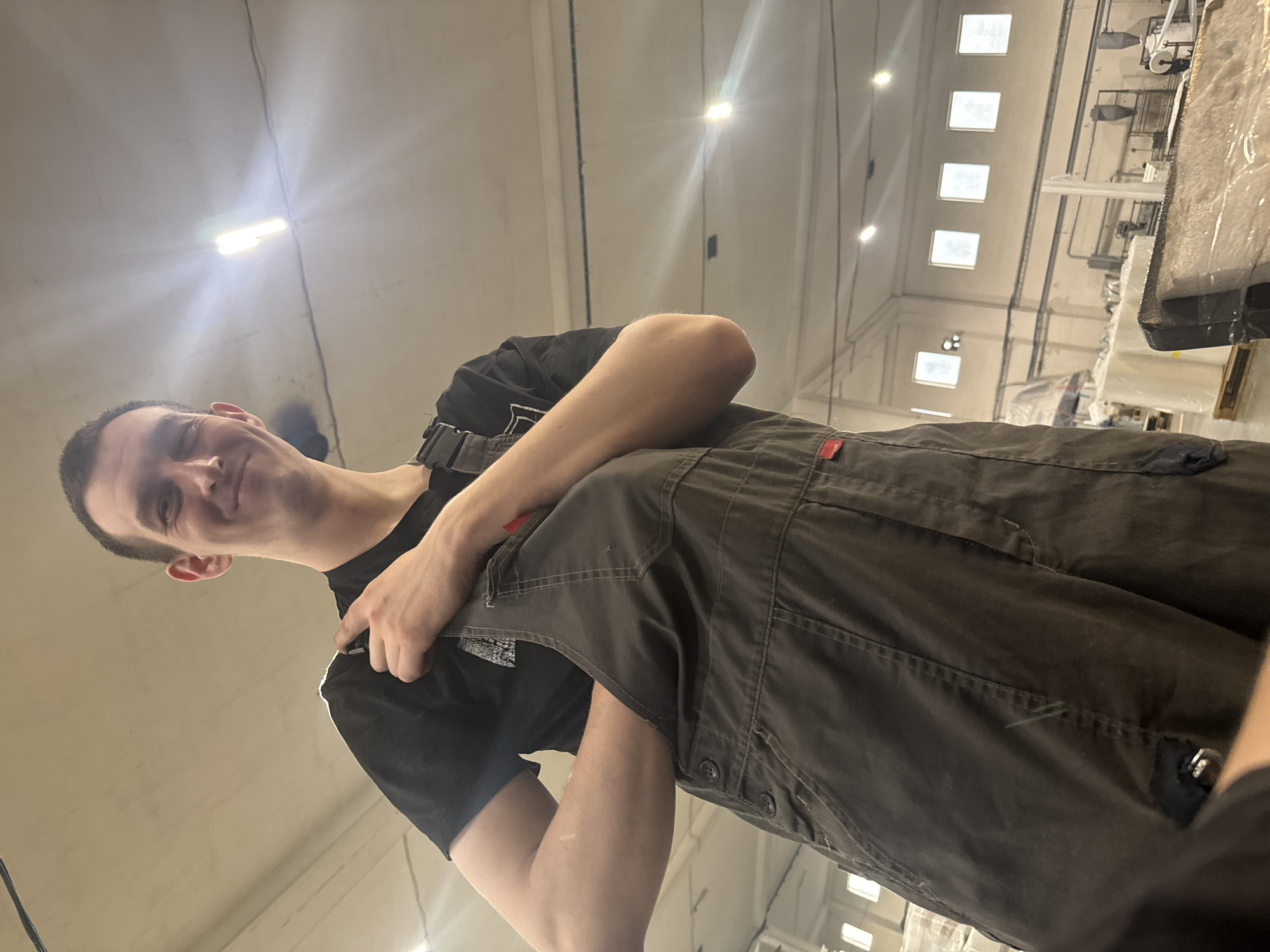 A male individual is depicted from the chest up in a large industrial facility located in Dikaya Gar', Russia. He has short dark hair, a clean-shaven face, and a light complexion, exhibiting a slight smile or smirk as he looks upward and towards the viewer's left. He is wearing a black t-shirt under dark grey overalls. His left arm is bent at the elbow with his hand positioned inside a front pocket of the overalls, while his right hand grips the right shoulder strap of the overalls. A small red tag is visible on the lower left side of his overalls. The background reveals a vast, light-colored ceiling with numerous industrial light fixtures, including a prominent, intensely bright elongated fluorescent light and several smaller, square lights. In the distance, multiple square windows are arranged high on a wall, admitting ambient light. Industrial equipment, including machinery and stacks of white rolled material on pallets, occupies the mid-ground. A rectangular object, wrapped in clear plastic and showing a textured, possibly fibrous or foamy interior, is partially visible in the lower right foreground.