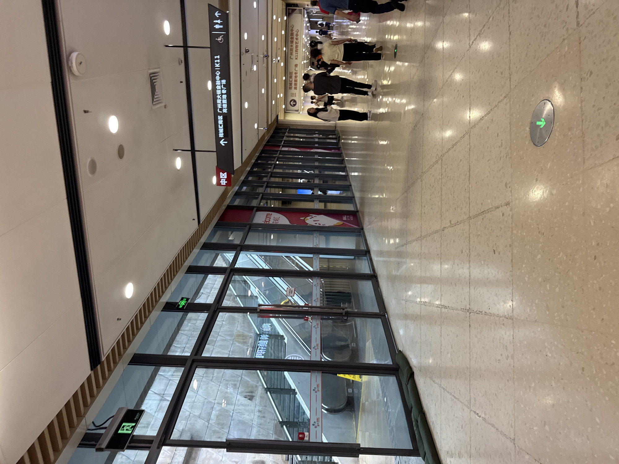 This is a medium shot of an indoor pedestrian walkway with people walking and standing. The walkway is covered with light-colored tiled flooring. Along the left side of the walkway, there is a long stretch of glass-fronted retail spaces, behind which escalators and stairs are visible. Some of the glass panels have red and white striped safety tape and additional signage. Hanging from the ceiling are multiple light fixtures and directional signs. One prominent sign, hanging above the walkway, indicates directions to "Huacheng Nan District," "Guangzhou Daming Financial Center K11," and "Gaode Land Square." Another sign with a green arrow on a circular metal plate is embedded in the floor. People in the image are dressed in casual attire, and their actions suggest normal pedestrian movement within a public space. The overall environment appears to be a modern commercial or transportation hub. The image is likely taken in Tianhe District, China, based on the directional signage.