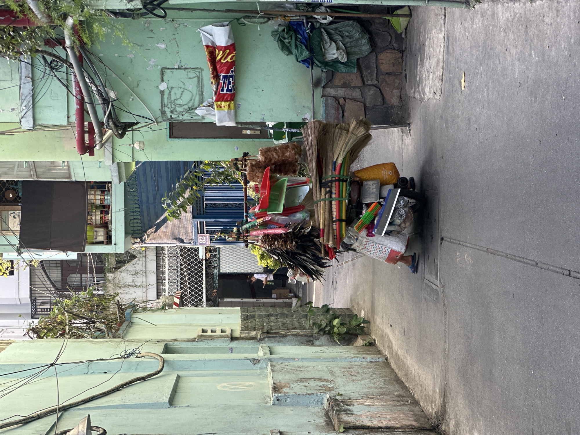 The image depicts a narrow, weathered urban alleyway, consistent with residential areas in Thủ Đức, Vietnam, during daytime. A motorized two-wheeled vehicle, heavily laden with household cleaning products, occupies the center of the alley, moving away from the viewer. The vehicle is piled high with brooms, feather dusters, plastic dustpans, and various bundled goods. A person's lower legs and feet, clad in blue flip-flops and a patterned skirt, are visible, indicating they are operating the vehicle.
The alley is flanked by two-story buildings, predominantly painted in a faded mint green, showing significant weathering, peeling paint, and exposed concrete patches. Both walls feature an extensive, dense network of black electrical wires and conduits, with some red and grey pipes visible on the right-hand building. Small green plants grow from the bases of the walls.
On the right side of the alley, near the foreground, the building's base transitions to a rough stone or concrete foundation. A small brown door is visible further along this wall. Below the door, bundles of green and white fabric or plastic bags are accumulated. A red and white banner, possibly commercial packaging, is affixed to the wall above these items.
Further down the alley, in the background, another individual in a white shirt and dark pants stands near an open doorway with a diamond-patterned metal gate. This person appears to be looking down at an object held in their hands. The building number "82" is visible on a small sign next to this doorway.
The ground is paved with dark grey asphalt or concrete, exhibiting minor cracks and variations in texture. Overhead, a dense network of tangled electrical and communication cables crisscrosses the alley between the buildings.