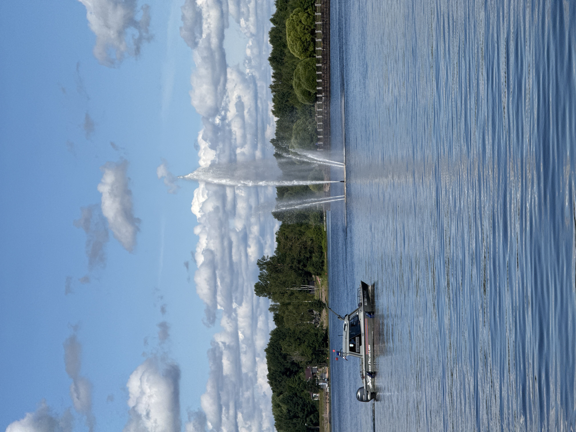 In Vyborg, Russia, a boat with the registration "AA 5001 RU547" cruises across a wide, blue body of water under a partly cloudy sky. The boat is dark gray and has a Yamaha outboard motor. In the background, a large fountain shoots a tall stream of water high into the air, with several smaller streams fanning out around it. Beyond the fountain, a dense line of lush green trees forms the shoreline. Further back, a few buildings are visible nestled among the trees on the left side of the image. A stone embankment with decorative elements runs along the water's edge in front of the trees, to the right of the fountain. The water's surface is rippled, reflecting the blue sky and white clouds.
