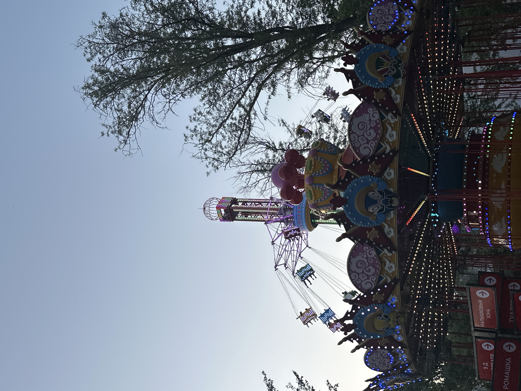 This is a photograph of an amusement park ride in Manas, Kyrgyzstan. The ride appears to be a carousel-style swing ride, with colorful lights and decorative elements. In the background, a tall tower with lights is visible, as well as several swings in motion with people riding them. The sky is clear and blue, suggesting it is daytime. Trees are visible in the background, indicating an outdoor setting. Signs with Russian text are present in the foreground, with one reading "МАЛЫШКОВЫЙ ЗОНА" which translates to "KIDS ZONE." The overall scene depicts a lively and festive atmosphere characteristic of an amusement park.