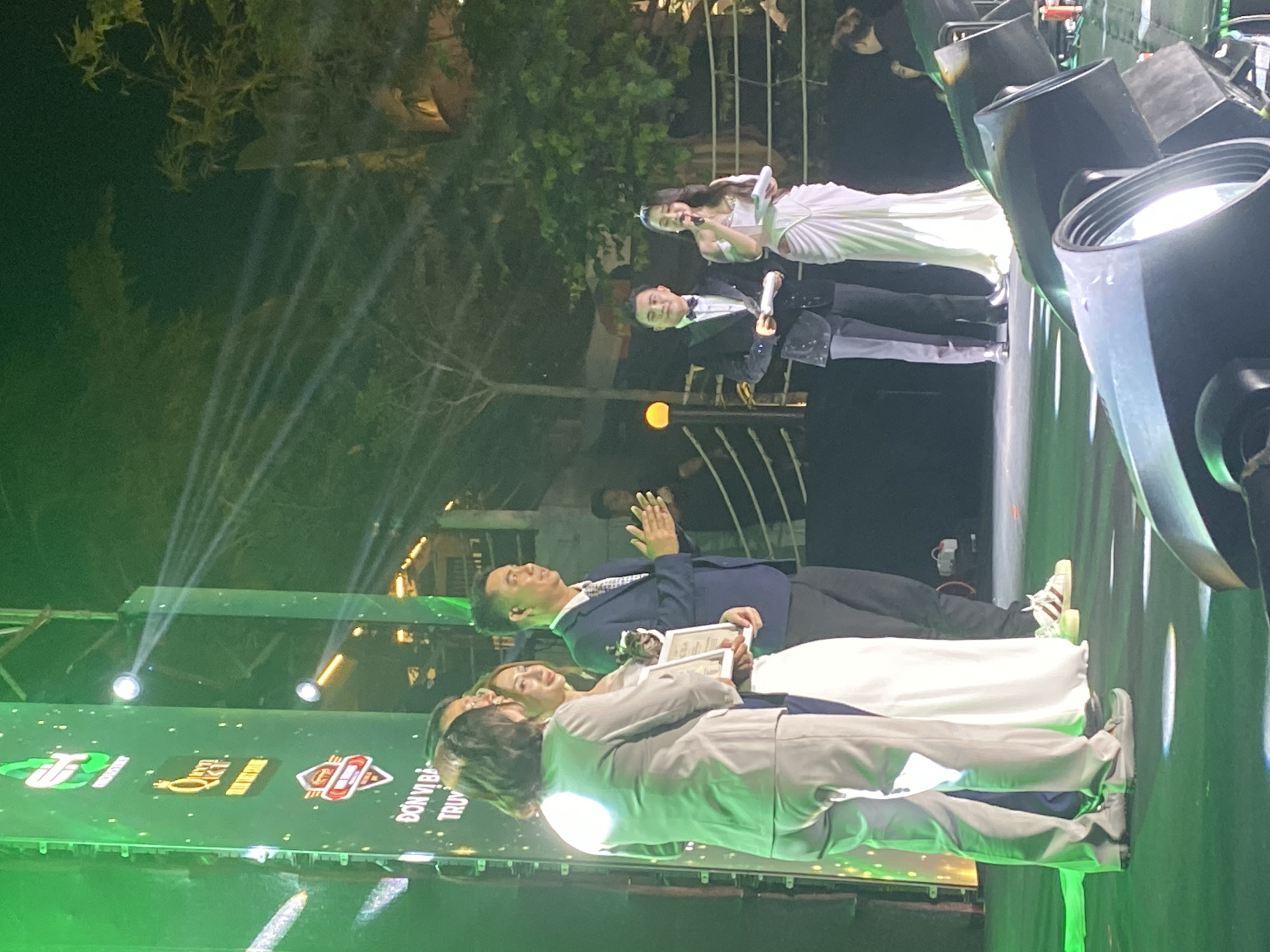 In Mũi Né, Vietnam, a group of individuals are on a stage, illuminated by bright green lights. A large vertical screen displaying logos and text in Vietnamese is to the left of the frame. Several people are gathered near the center, with some appearing to be receiving or presenting awards. One woman in a white dress is holding a framed award. To the right, a man in a tuxedo and a woman in a white gown are holding papers and speaking into microphones. The background shows trees and structures, suggesting an outdoor evening event. The stage floor is a dark green, with spotlights creating bright circular reflections.