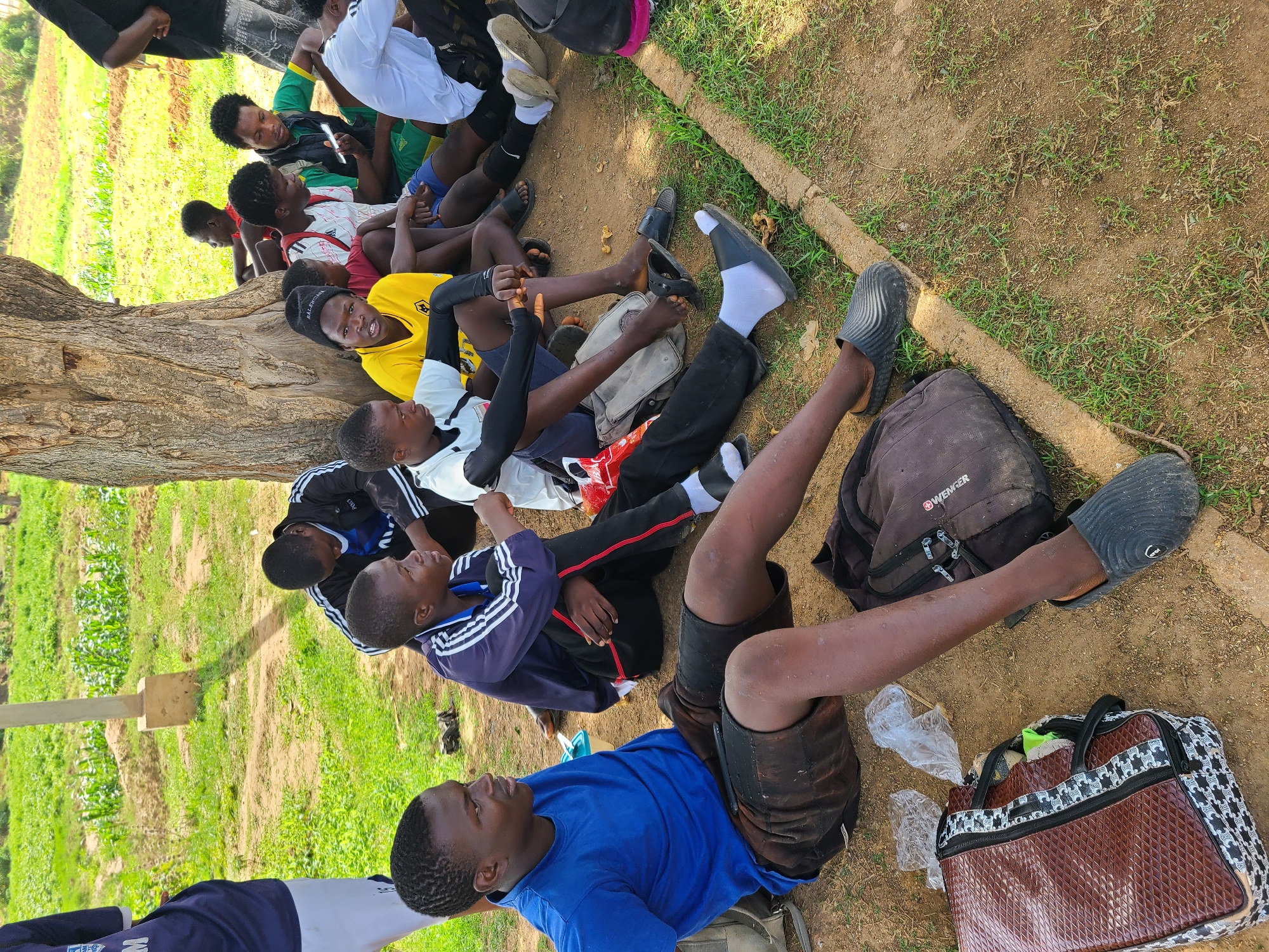 A ground-level shot depicts a group of young men, predominantly Black, resting beneath the shade of a tree. The men are dressed in an assortment of athletic wear, including jerseys in colors such as blue, yellow, red, white, and green, with some wearing casual attire like t-shirts and tracksuits. They are lying on the ground, which consists of dirt and sparse grass.
To the right of the frame, near one of the men, are a backpack and a brown and black patterned duffel bag. Some of the men are interacting, while others are looking at their phones. The scene is set outdoors, with green vegetation visible in the background. The location is Unknown, Unknown.