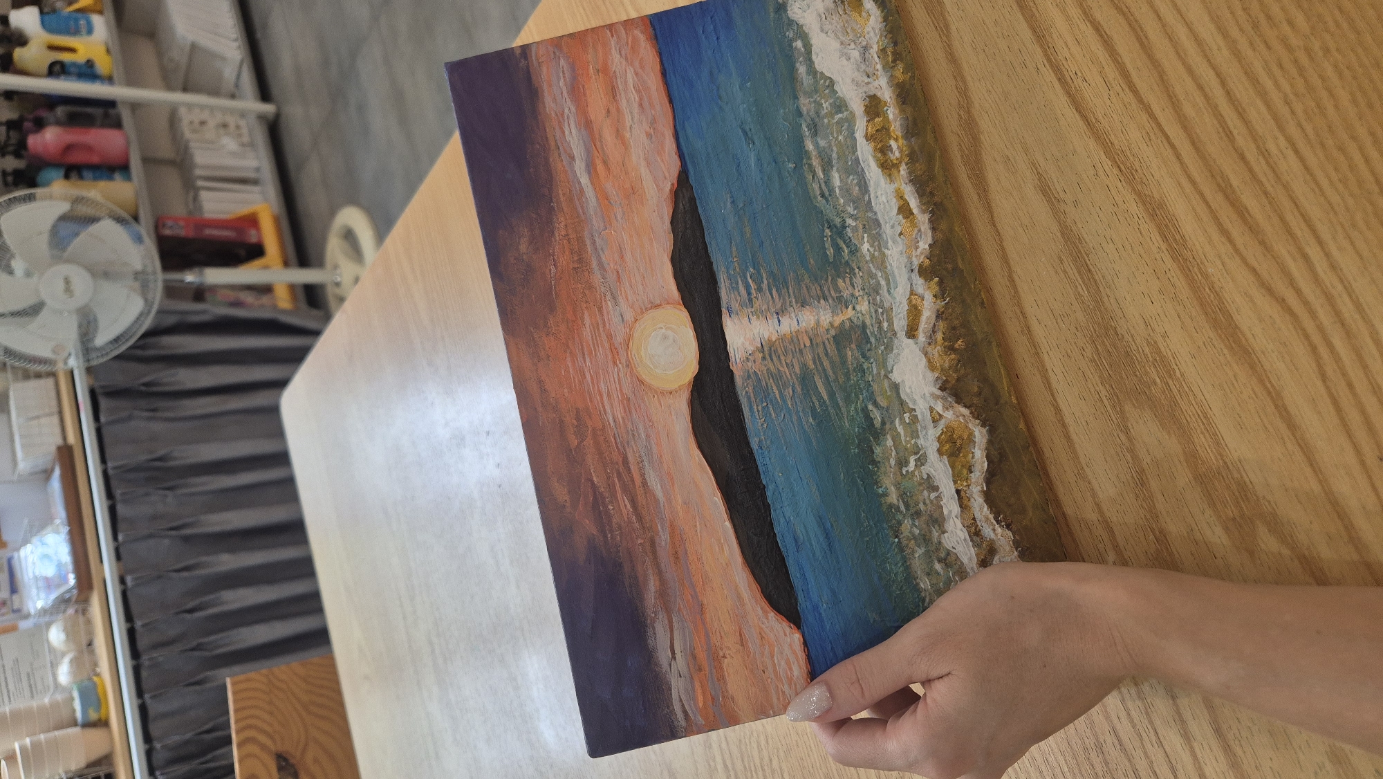 A person's left hand with glittery fingernails is holding a rectangular acrylic painting of a sunset over the ocean. The painting depicts a dark silhouette of a mountain range against a vibrant orange and purple sky. The sun, a bright yellow circle, is partially obscured by the mountains, casting a reflection on the turquoise water. The foreground of the painting shows white waves crashing onto a golden-brown shore. The painting is placed on a light-colored wooden table. In the background, shelves with various items, including bottles and books, are visible, along with a white oscillating fan. The scene does not provide specific information that can be used to determine a location in the city of Da Nang, Vietnam.