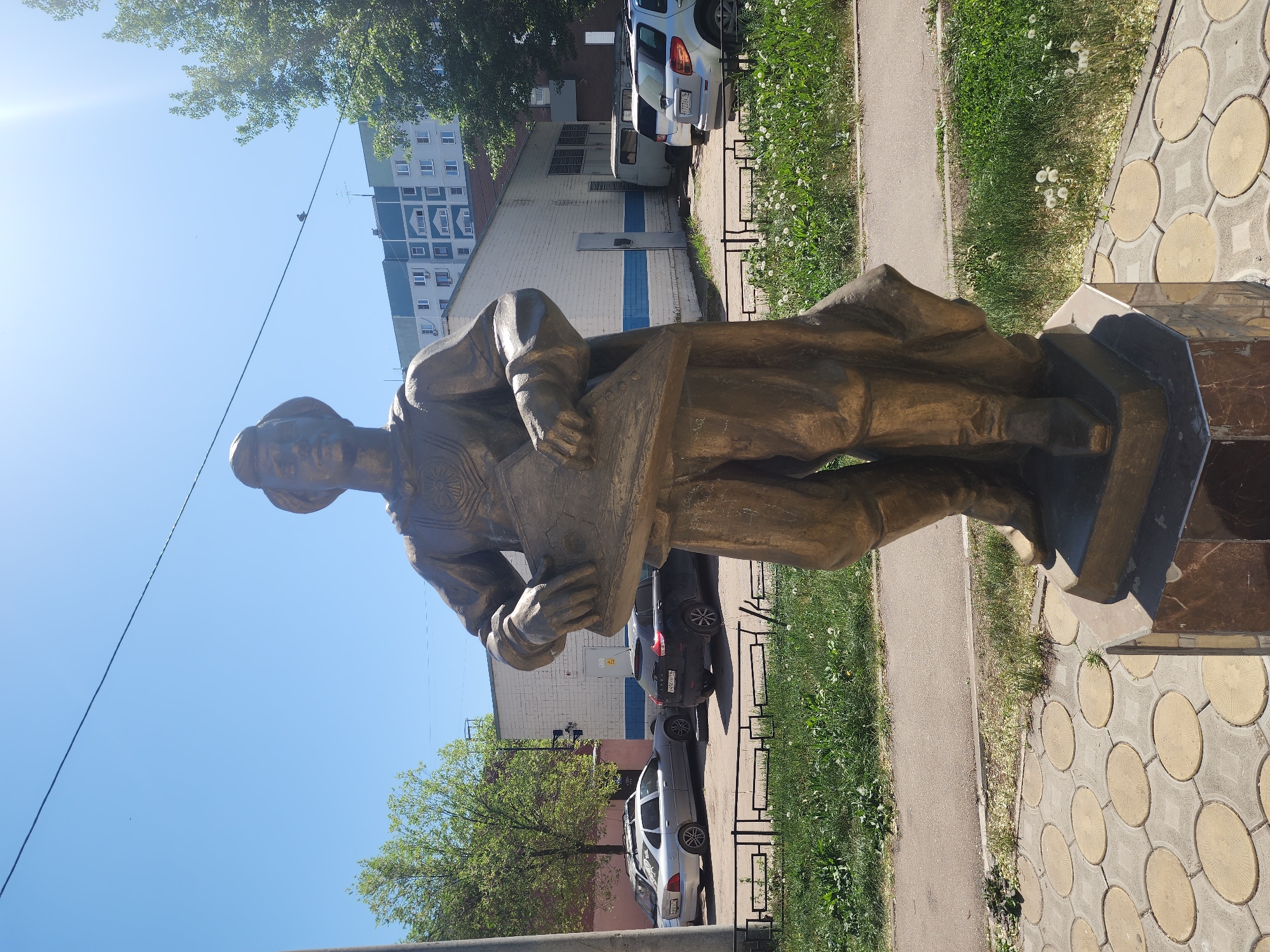 The image depicts a bronze statue of a man in Kazan, Russia. The statue stands on a dark base, placed on a light grey and yellow patterned pavement next to a sidewalk. The man is wearing a coat or robe and appears to be holding a tablet or scroll in his hands.
Behind the statue, a grassy area is visible next to the sidewalk, followed by a black metal fence. Beyond the fence, several cars are parked along the street. Behind the cars, there is a light-colored building with blue accents, and a tall building is visible in the background. Trees surround the buildings. The sky is clear and blue.