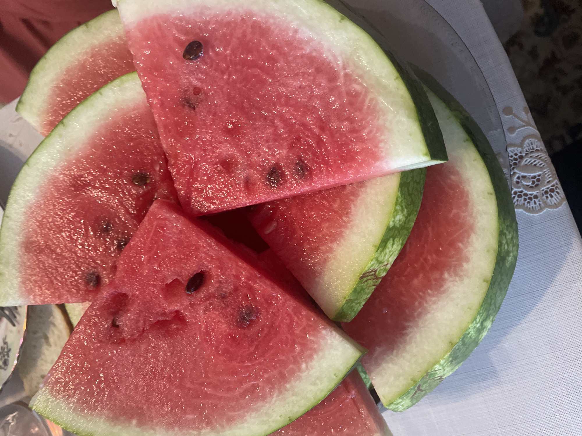 A pile of sliced watermelons is displayed on a white tablecloth. The watermelons are cut into triangular wedges, revealing their bright red flesh and scattered black seeds. The rind is a mix of green and white. The scene is lit from above, casting shadows on the tablecloth. The location is identified as Brest, Belarus.