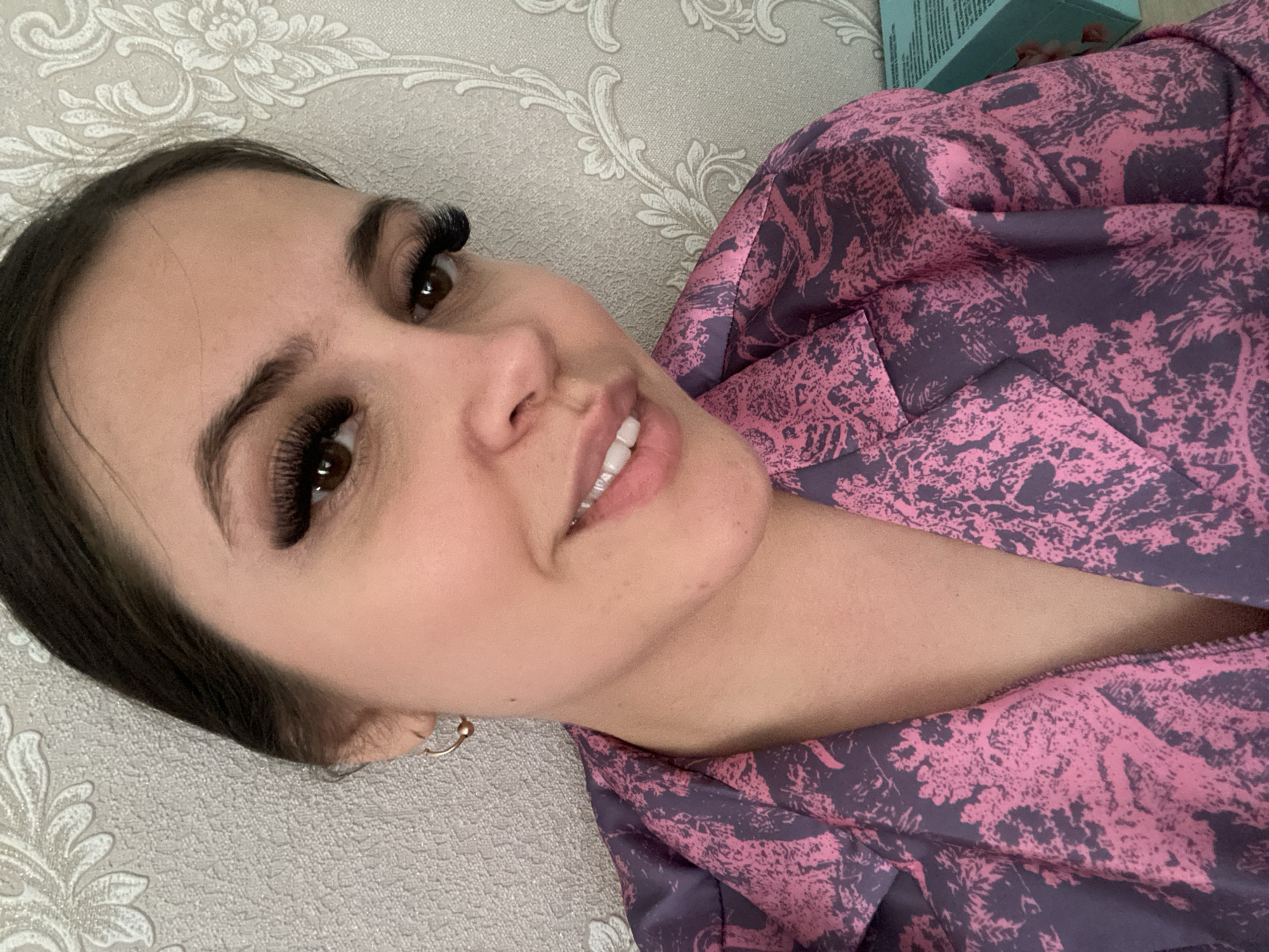 This is a close-up indoor portrait of a young woman, likely in her twenties or thirties, taken at an angle as if she is reclining. She has dark hair, dark brown eyes, and striking, voluminous false eyelashes. Her eyebrows are dark and well-defined. She is looking towards the camera with a subtle, pleasant smile, revealing her neat, white teeth. A small, gold-colored hoop earring is visible in her left ear.
The woman is wearing a garment with a distinctive abstract or stylized floral pattern in shades of light pink and dark purple, possibly a robe or a top. The background is a light-colored wall, likely wallpaper, with a textured damask or floral pattern in a slightly darker shade. In the upper right corner, a small, out-of-focus light blue box or object is partially visible. No discernible text is visible on any objects. The overall impression is a casual, intimate portrait focusing on her facial features. The image is set in Chaikovsky, Russia.