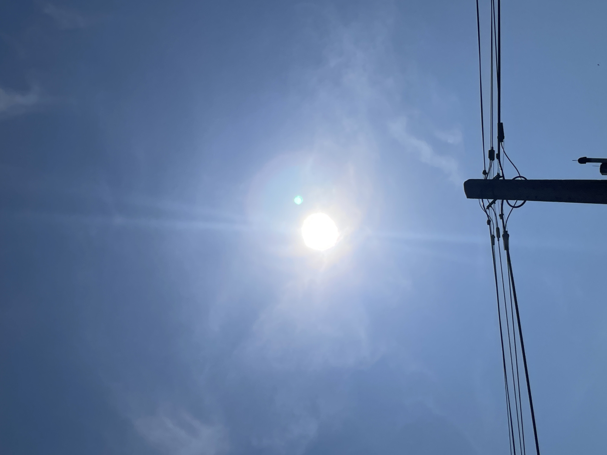 An outdoor, eye-level, vertical shot captures a bright, sunlit sky. The sun is visible as a bright, circular object in the upper center of the frame, emitting a sunburst effect. Thin, wispy clouds are scattered across the expanse of the blue sky. In the foreground, the upper portion of a dark, silhouetted telephone pole is visible, supporting multiple dark wires that stretch horizontally across the frame. The pole and wires are in sharp focus, contrasting with the softer focus of the sky. The scene is set against a clear, blue sky, suggesting a bright, sunny day. There are no people or animals visible in the image. The overall impression is one of a bright, clear day with an overhead view of utility infrastructure.