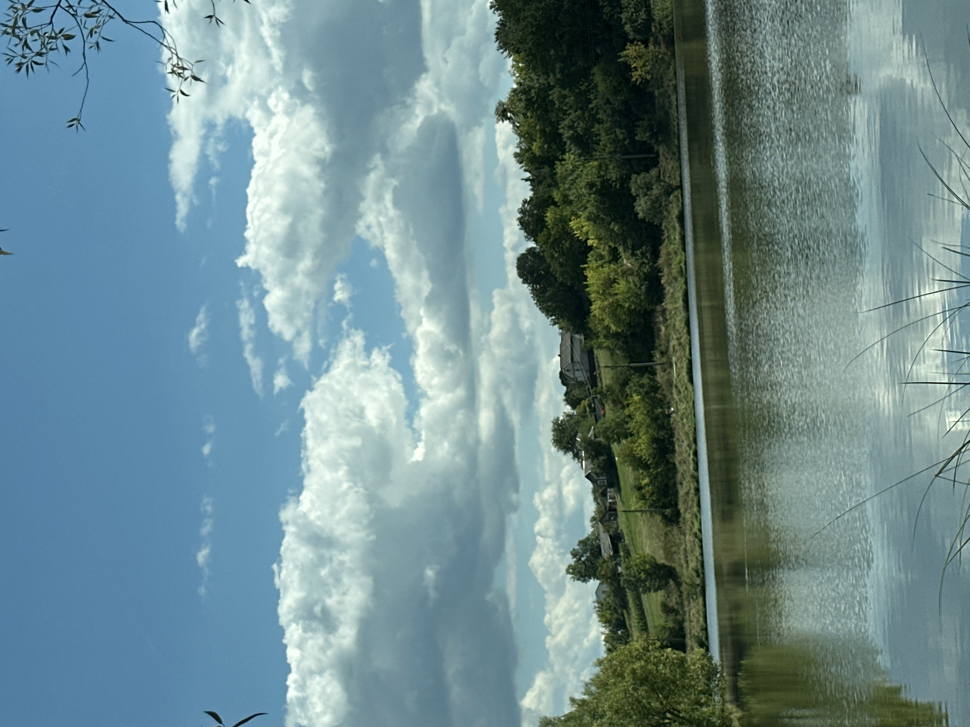 The image depicts a serene lake scene in Makiv, Ukraine, under a partly cloudy sky. The foreground features the calm surface of the lake, reflecting the sky and surrounding greenery. In the mid-ground, a gently sloping hillside is covered with lush green trees and some scattered houses. The houses appear to be rustic and integrated into the natural landscape. Tall grass and reeds are visible at the edge of the water in the lower foreground. The sky is a bright blue, dotted with fluffy white and gray clouds, indicating fair weather with some cloud cover. The overall impression is one of a peaceful rural environment.