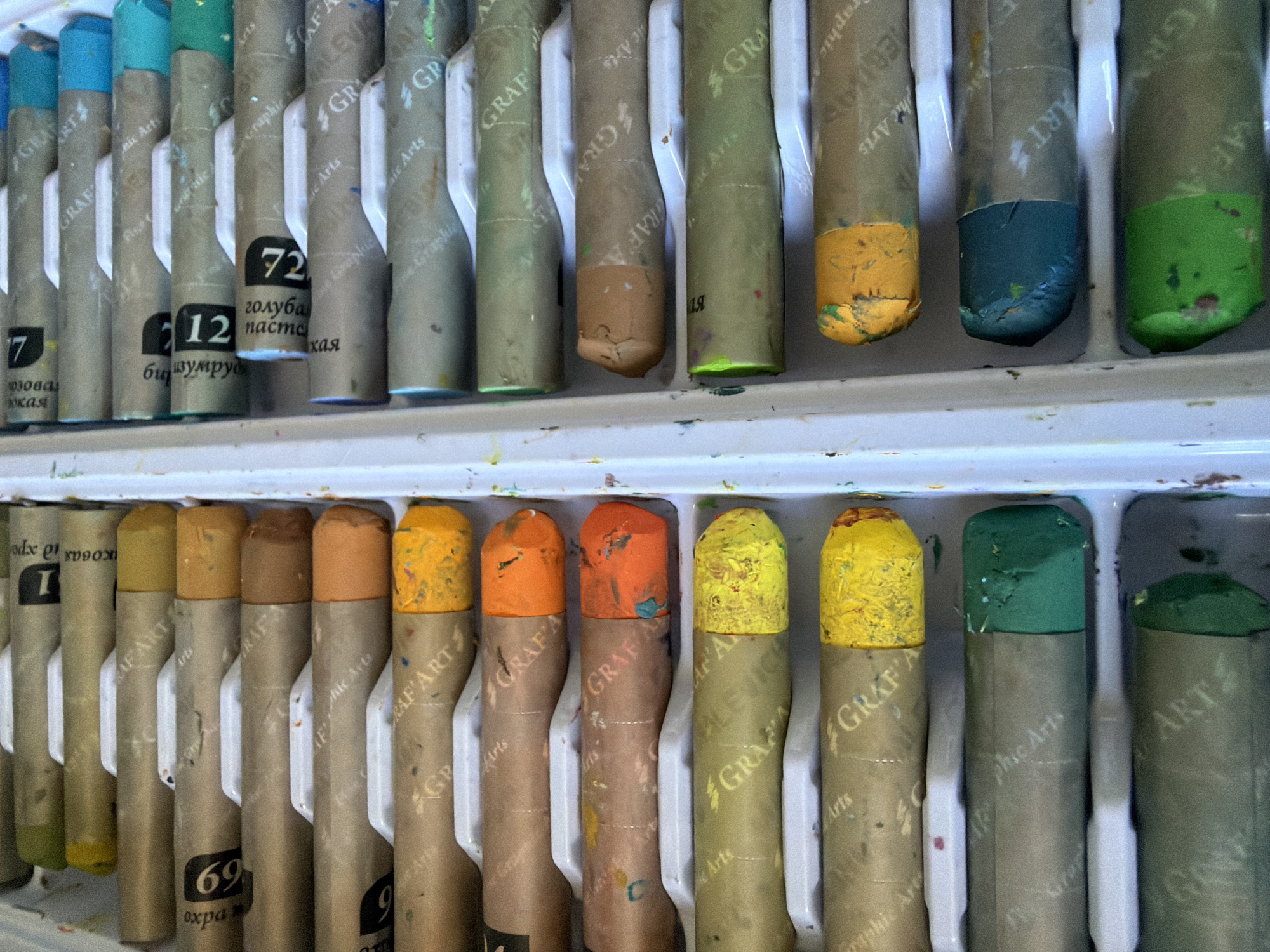 The image shows a close-up, top-down view of an open box of oil pastels. The pastels are arranged in rows within a white plastic tray. The wrappers of the pastels, made of brown paper, are visible. Many of the wrappers have text and logos, including "GRAF ART". The tips of the pastels, showing their colors, are also visible. The colors include various shades of yellow, orange, brown, green, and blue. Some of the pastel tips are chipped or worn, indicating use. The background is white, consistent with the interior of a plastic box. There are no people or other objects in the image. The scene appears to be an indoor setting, with no indication of a specific location in Ufa, Russia, or any other city.