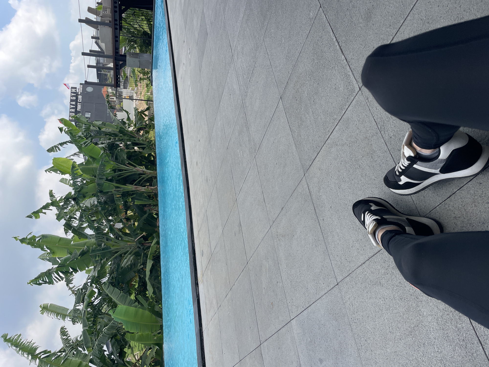 The image captures a poolside scene in Mengwi, Indonesia. In the foreground, a person's legs, clad in black leggings and athletic shoes with white laces, are visible, suggesting a moment of rest or relaxation. The ground is paved with large, light gray square tiles. Beyond the tiled area lies a vibrant blue swimming pool, its surface gently rippling. Behind the pool, a dense cluster of lush green banana trees forms a natural backdrop. In the distance, a multi-story building with a sign that reads "SURYA GYM FIGHT CLUB" is visible, topped with a small Indonesian flag. The sky above is a mix of blue and white clouds, indicating a bright, possibly warm day. The overall impression is one of an outdoor leisure space, possibly within a resort or fitness facility, set against a tropical environment.