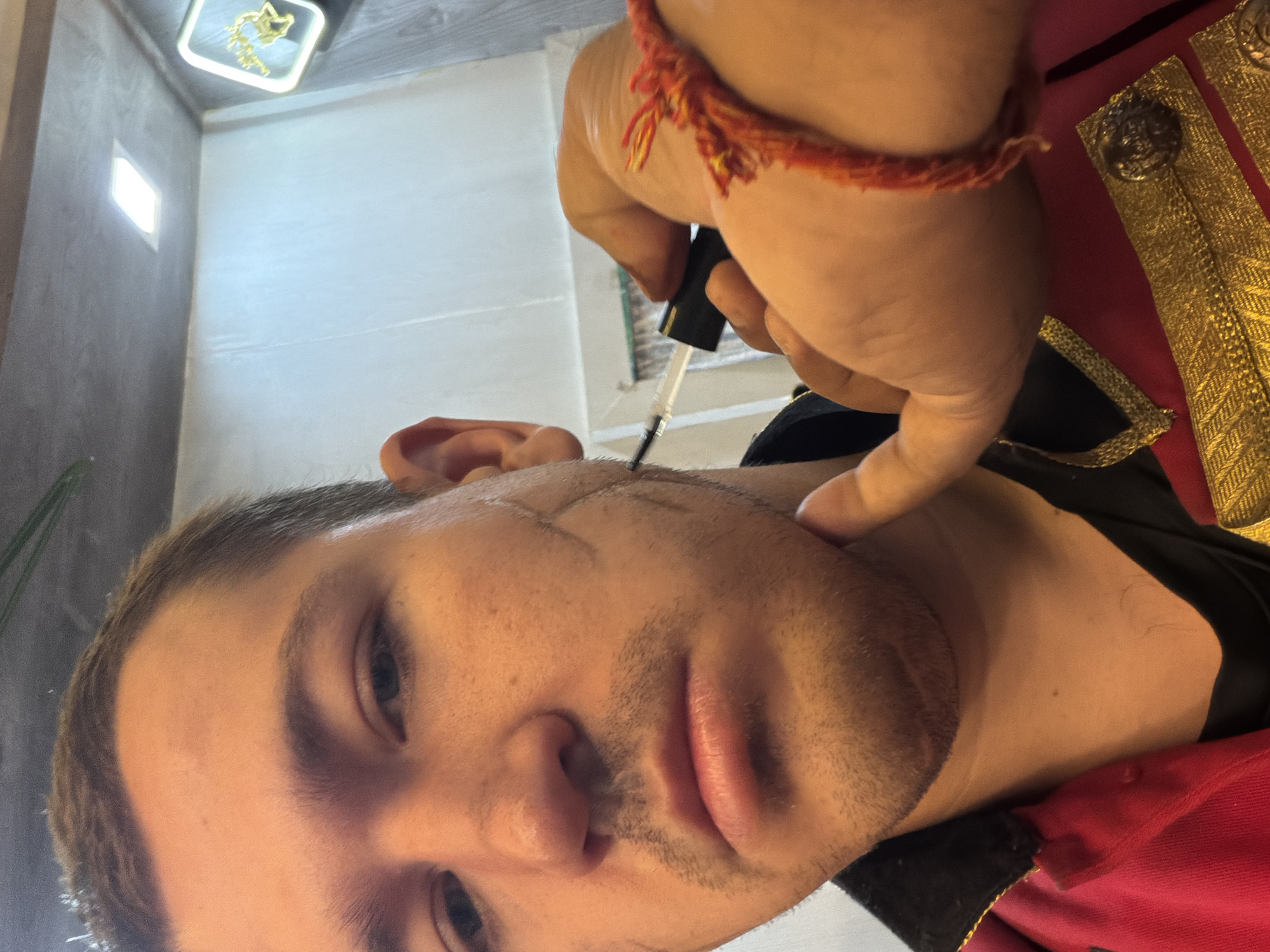 The image shows a close-up of a person's face, with their right hand applying makeup to their cheek. The person has short, brown hair, and appears to be wearing a red jacket with gold trim. The background is blurred but suggests an indoor setting. The image does not contain any discernible landmarks or features that would indicate its location in Hyderabad, India, or any other specific city.