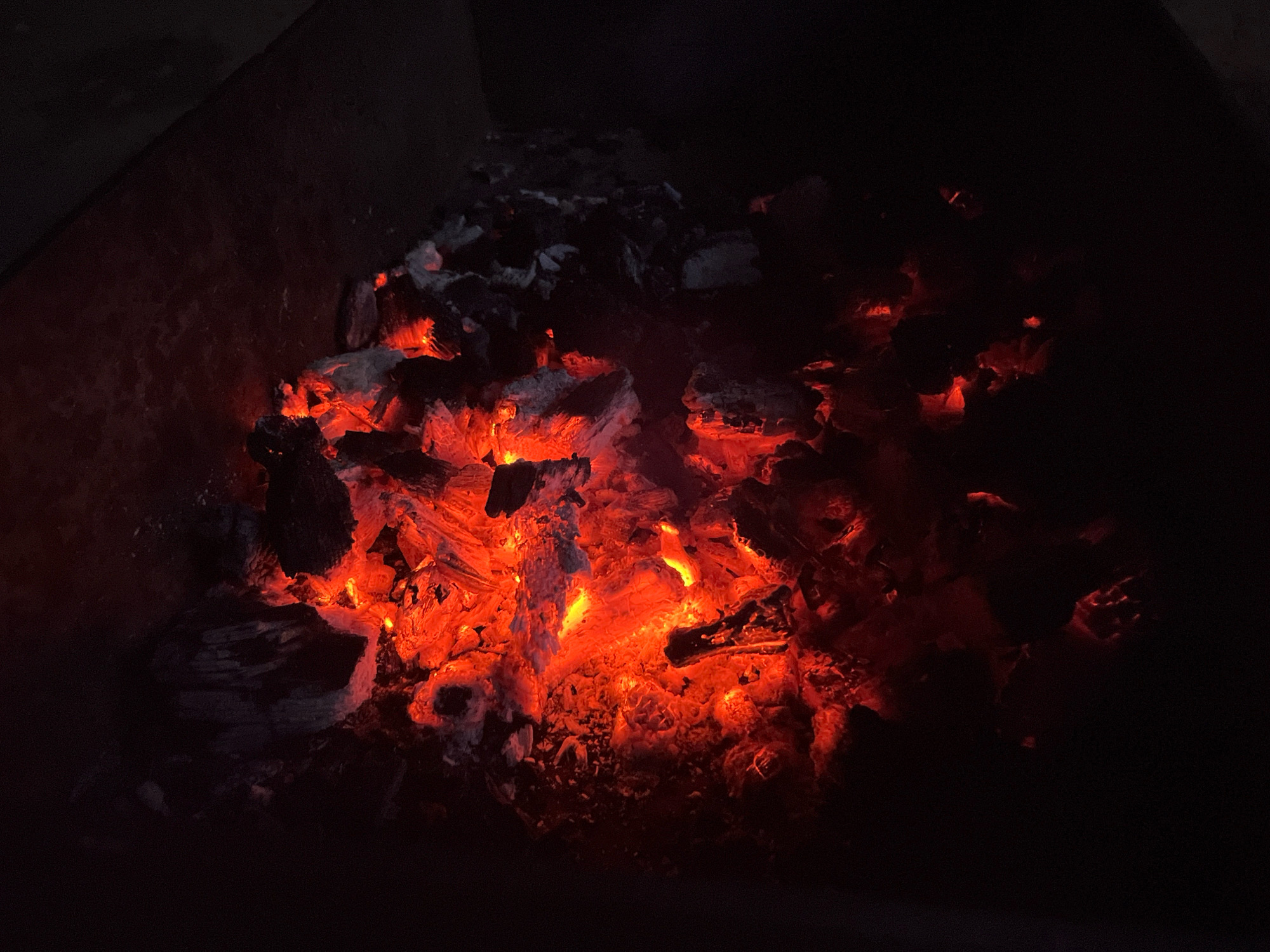 The image shows a close-up view of glowing red-hot embers and charcoal in a dark environment, possibly a barbecue pit or fireplace. The coals are clustered together, with some appearing brighter than others, indicating varying temperatures. Ash and partially burnt wood fragments are visible amidst the glowing embers. The background is predominantly black, with hints of dark, textured surfaces on the left, suggesting the confines of a container. The scene is dimly lit, with the primary light source being the intense red and orange glow of the coals. There are no people, animals, or specific landmarks visible that would indicate a precise location within Taganrog City District, Russia. The focus is solely on the fire and its remnants.