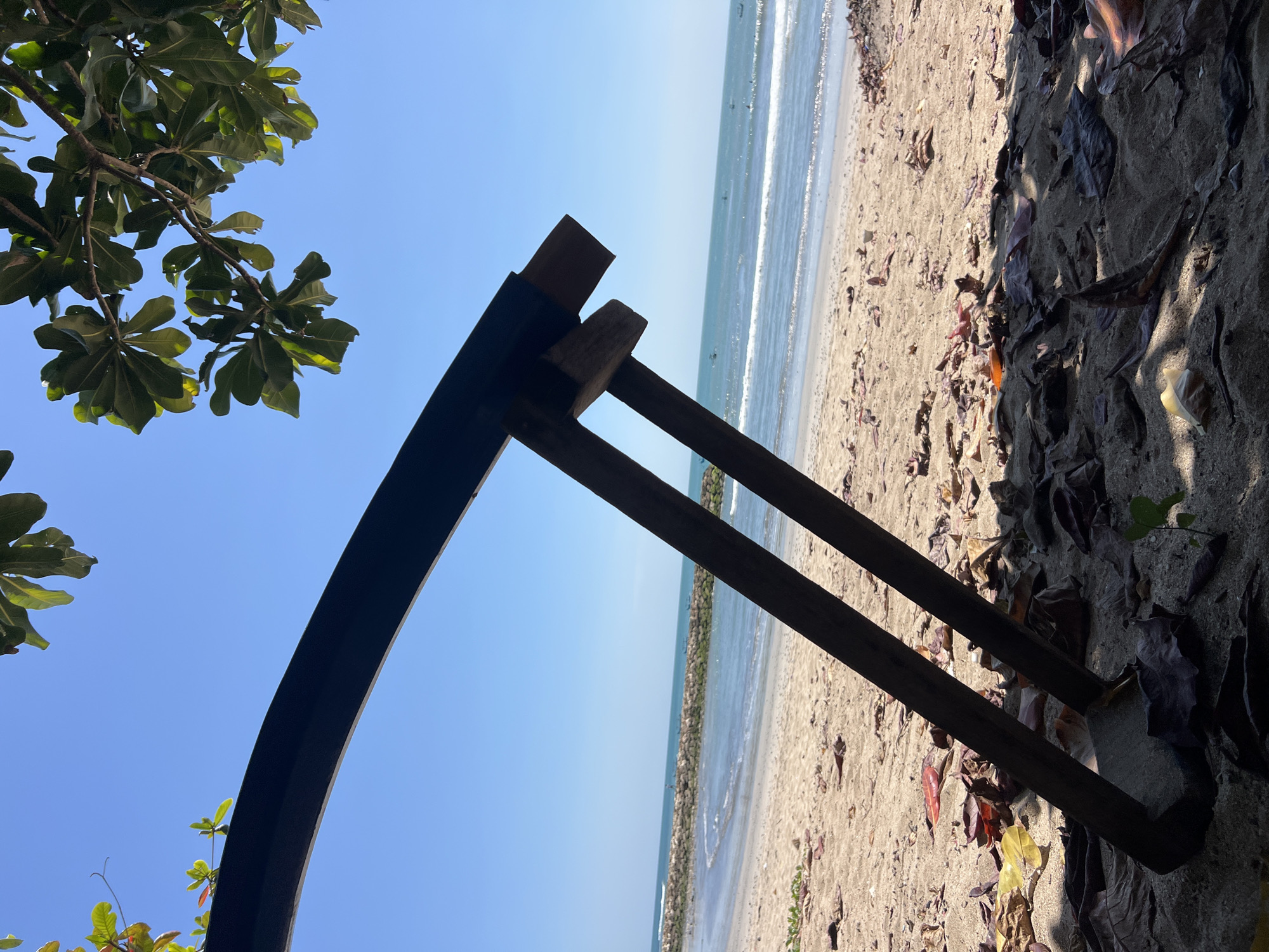 The image shows a close-up view of a rustic wooden structure, possibly a part of a beach umbrella or shelter, in the foreground. Two dark wooden posts angle upwards from the sandy ground, which is littered with fallen brown and red leaves. Beyond the structure, a tranquil beach scene unfolds under a clear blue sky. The sand transitions to the gentle waves of the ocean, with a stone breakwater visible in the distance. Small boats are scattered across the water. Green foliage from a tree hangs into the frame from the top, casting a shadow on the sand. The overall impression is one of a peaceful, sun-drenched day at the beach in Kuta, Indonesia.