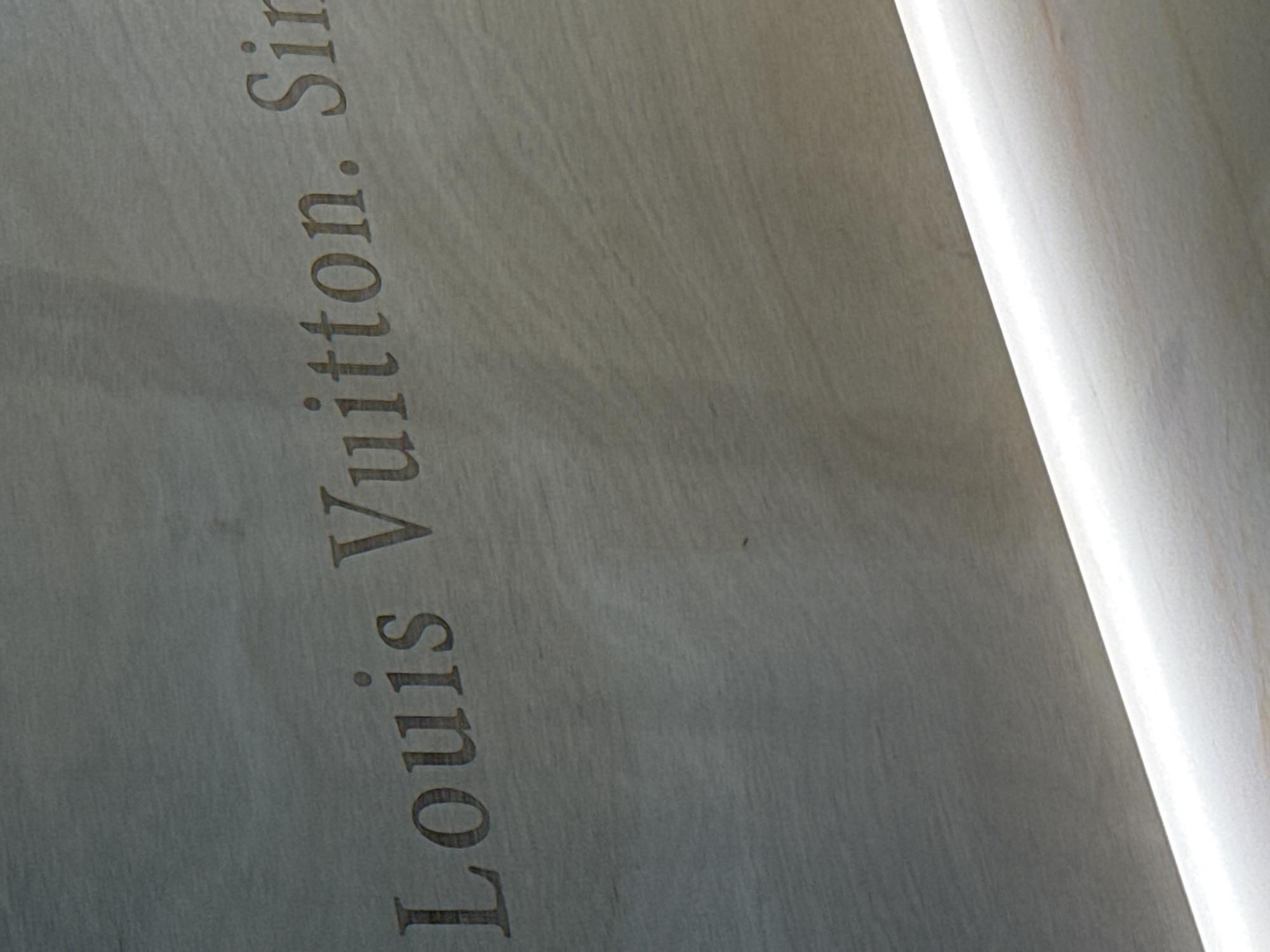 A close-up, slightly angled shot shows the words "Louis Vuitton. Sin." etched into a textured, light-colored surface. The text is in an elegant, serif font and is darker than the background. A bright, white light source runs diagonally across the bottom of the frame, illuminating a portion of the surface and creating a gradient effect. The overall impression is of a branded inscription on a material like wood or stone. The location in Nong Prue Subdistrict, Thailand, is not discernible from the image itself.