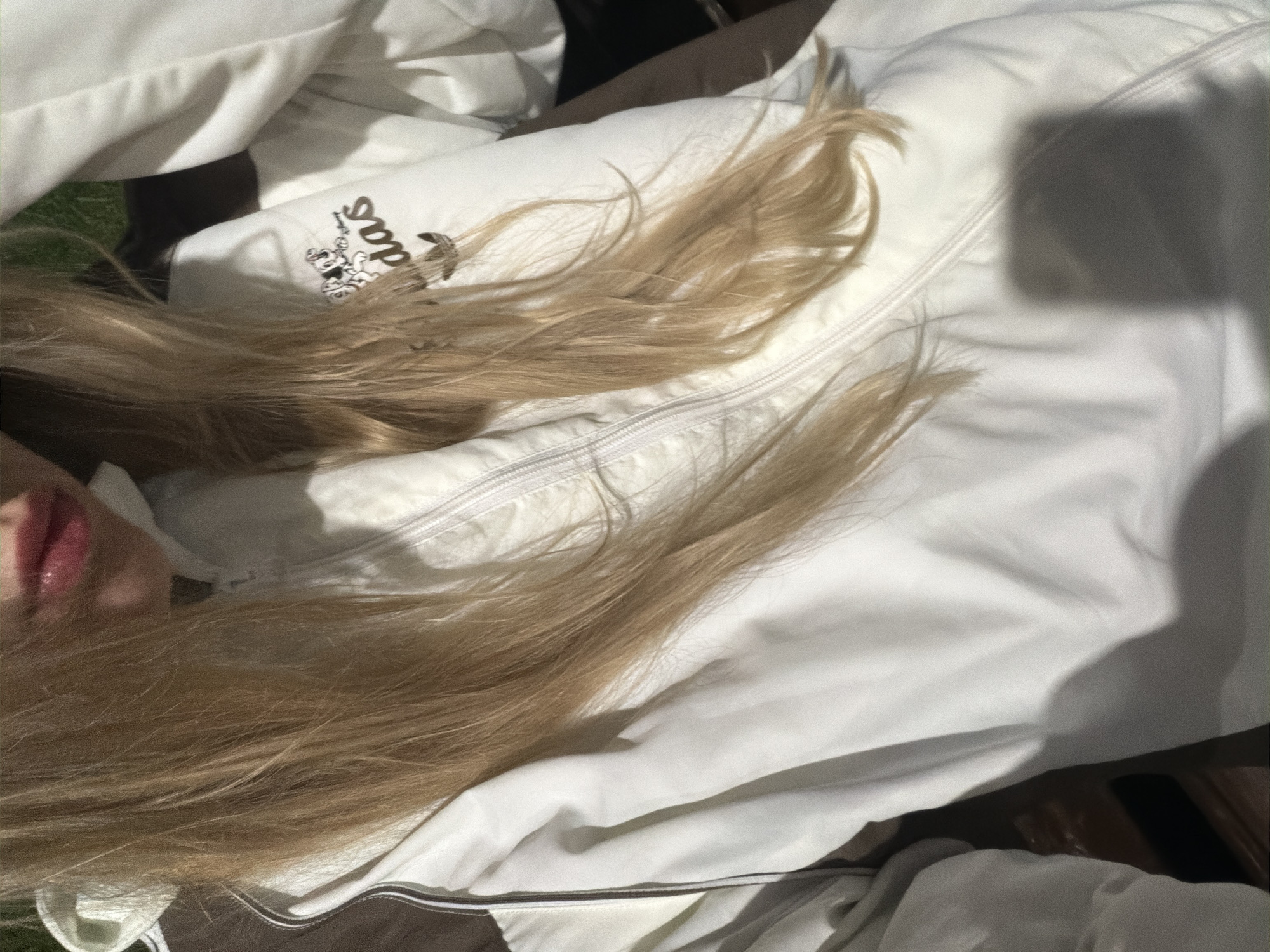 An overhead shot shows a person lying on a white jacket. The jacket has a zipper and an Adidas logo on the left side. The person has long, blonde hair that is spread across the jacket. A portion of the person's face, including their lips, is visible. The person is outdoors, possibly on a bench or similar surface. The location is unknown.