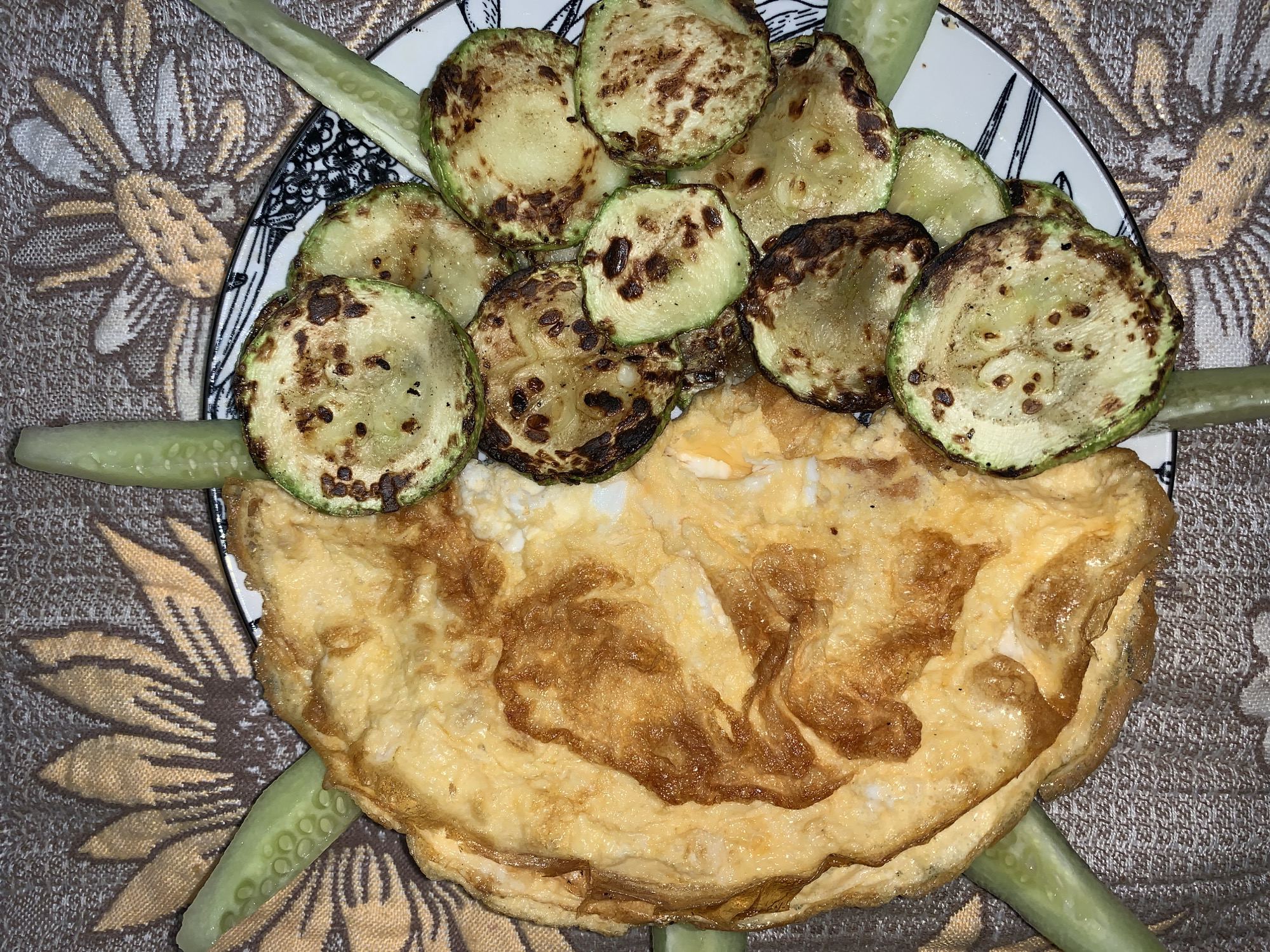 The image displays a close-up, top-down view of a prepared meal served on a plate, set against a patterned fabric background. The central subjects are a golden-brown omelette, a pile of grilled zucchini slices, and several cucumber sticks.
The omelette, positioned towards the bottom of the plate, is folded and exhibits a rich, toasted golden-brown color with darker, caramelized patches, suggesting a well-cooked exterior. Resting on top of the omelette is an arrangement of sliced zucchini. These zucchini rounds are light green with noticeable dark brown to black char marks, indicating they have been grilled or pan-fried. Thin, light green cucumber sticks are placed around the edge of the plate, partially beneath the main dishes, serving as a fresh garnish or side.
The food is presented on a white plate with a distinct black and white abstract or geometric pattern adorning its rim. This plate rests on a textured fabric surface, likely a tablecloth or placemat. The fabric features a beige or light brown background color, overlaid with a prominent, lighter-colored floral design that resembles a stylized sunflower. The pattern appears to be woven or embroidered.
The lighting is even and appears to be indoors, casting a slightly warm tone over the food. There is no visible text in the image. The overall impression is that of a freshly prepared and aesthetically appealing meal, possibly breakfast or a light lunch.