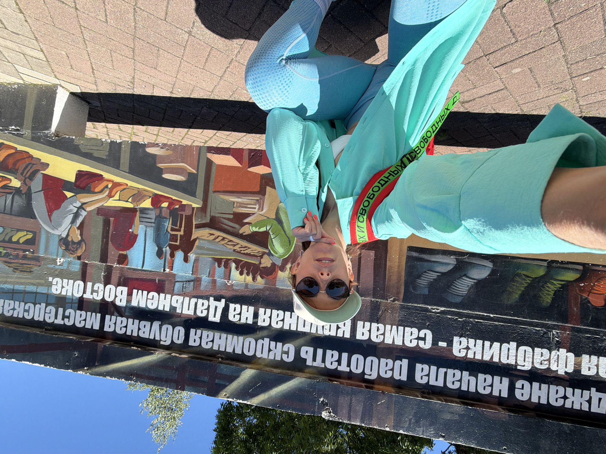 A woman wearing a light blue, long-sleeved open shirt over a white sports bra and blue leggings is posing for a selfie in front of a mural. The mural depicts a scene with text in Russian, along with illustrations of shoes and people working in a workshop. The text, as visible in the cropped images, reads "ДЖАНЕ НАЧАЛА РАБОТАТЬ СКРОМНАЯ ОБУВНАЯ МАСТЕРСКАЯ НА ДАЛЬНЕМ ВОСТОКЕ" which translates to "Dzhan began to work, a modest shoe workshop in the Far East". The woman has blonde hair, wears sunglasses and a light green baseball cap. A red strap with black lettering, also in Russian, is visible across her chest. The ground is paved with reddish-brown bricks. The scene appears to be outdoors on a sunny day, with trees and a bright blue sky visible in the background. The mural is located in Birobidzhan, Russia, the administrative center of the Jewish Autonomous Oblast.