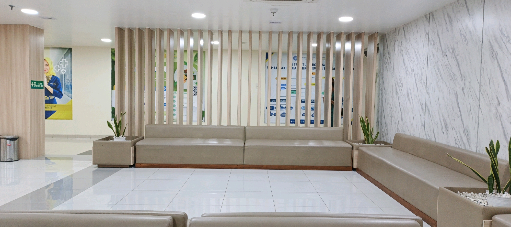 This image depicts a clean, modern waiting area within a facility in Trihanggo, Indonesia. The foreground features light-colored, tiled flooring with a subtle grey stripe pattern. A long, low-profile, taupe-colored sofa is the central object, situated behind a decorative wooden slatted partition. This partition adds a sense of privacy and aesthetic appeal.
To the left, a metal trash can stands near a wooden pillar. A large poster is visible on the pillar, featuring a woman in a yellow hijab and a blue uniform, along with some text and graphics. Another poster, with a more abstract design of circles and text, is partially visible behind the wooden slats.
In the middle ground, planters with small green snake plants (Sansevieria trifasciata) are placed beside the sofa. The background consists of a light-colored wall with a marble-like pattern. The overall impression is one of a well-maintained and organized space, likely a reception or waiting room in a clinic, office, or similar institution. There are no people actively engaged in any actions within the visible frame.
