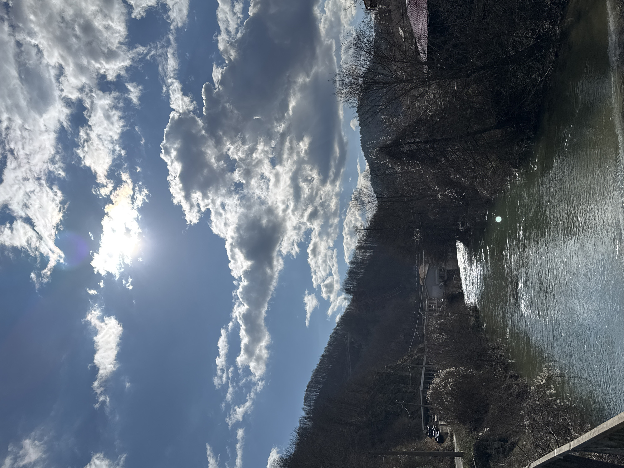 The image shows a view of Rudozem, Bulgaria, featuring a river reflecting sunlight. On the right is a body of water, reflecting a sunlit sheen. On the left is a cloud-filled sky. A hillside covered in bare trees is also visible. There are buildings on the riverbank. The overall impression is of a clear, bright day in a hilly region.