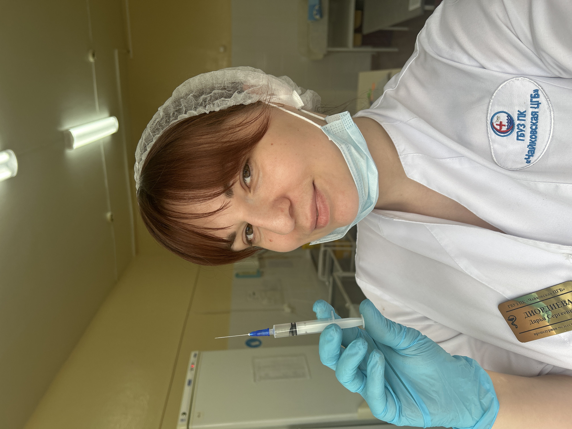 A female healthcare worker, presumably a nurse, is holding a syringe filled with a clear liquid. She is wearing a white medical coat, a disposable hairnet, and a light blue surgical mask pulled down beneath her nose. She has short reddish-brown hair and light skin. Blue latex gloves cover her hands. On her coat, a badge is visible with the text "ДИОРДИЕВА Дарья Сергеевы" (DIORDIEVA Daria Sergeevna) and below that, "процедурная медсест" (procedural nurse). Also visible is the logo of the hospital she works in: “Чайковская ЦГБ” (Chaikovskaya Central City Hospital). The location is Фоки, Russia. The background shows a medical setting with light-colored walls and a refrigerator.