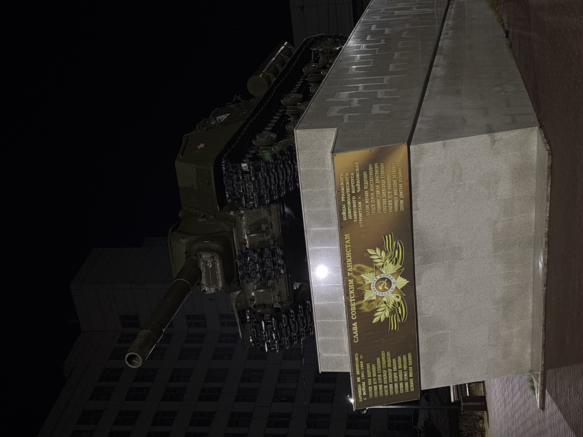 The image shows a Soviet tank monument at night in the city of Chaikovsky, Russia. The tank is mounted on a large concrete plinth. There is a plaque on the side of the plinth with an inscription that includes the text "СЛАВА СОВЕТСКИМ ТАНКИСТАМ" (Glory to Soviet Tankers) and lists names. A multi-story building is visible in the background. The scene is dimly lit.