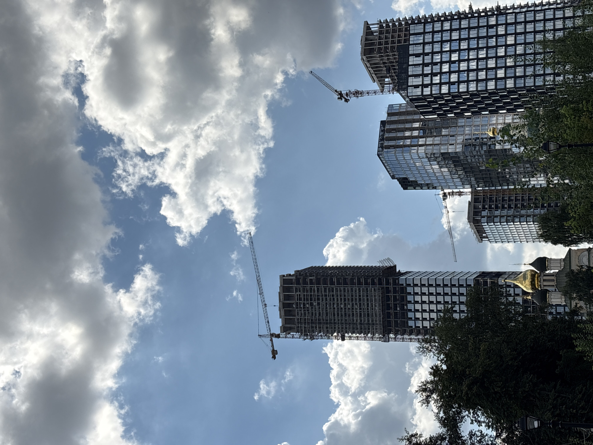 The image depicts a view of modern skyscrapers under construction in Moscow, Russia, contrasted with a traditional golden-domed church. The foreground features lush green trees and a lamppost. In the background, several high-rise buildings are in various stages of completion, with exposed scaffolding and cranes evident. One particularly tall skyscraper is prominently featured, its skeletal structure contrasting with the partially completed glass facades of neighboring buildings. The sky is a mixture of blue sky and dramatic, heavy clouds, suggesting a dynamic weather system. The golden domes of the church stand out against the urban landscape, symbolizing a blend of historical and contemporary architecture within the city.