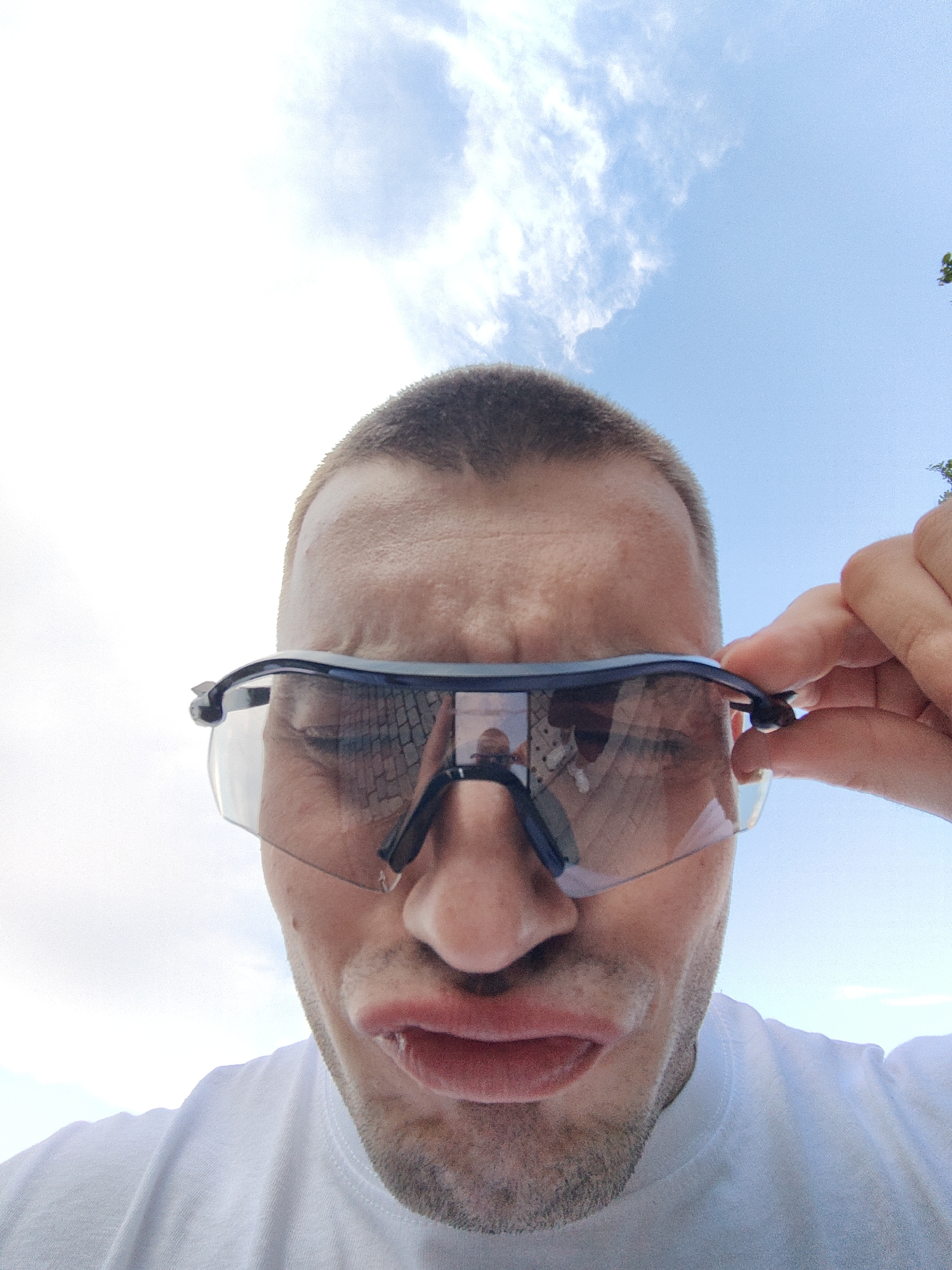 A low-angle, medium close-up shot depicts a light-skinned man with short, cropped hair, wearing sunglasses. He is looking down at the camera with a slight grimace or pout on his face. The sky is visible in the background, with a mix of blue and white clouds. The man is wearing a white t-shirt.
The sunglasses reflect a scene of a paved area, possibly a sidewalk or pedestrian zone, with a person or people standing on it. The man is holding the sunglasses with his right hand, with his index and thumb visible on the right side of the frame. The background suggests an outdoor setting. Based on the reflection in the sunglasses, it seems the image might be taken in a city. Since the location is specified as Shumen, Bulgaria, this is likely the city where the image was taken.