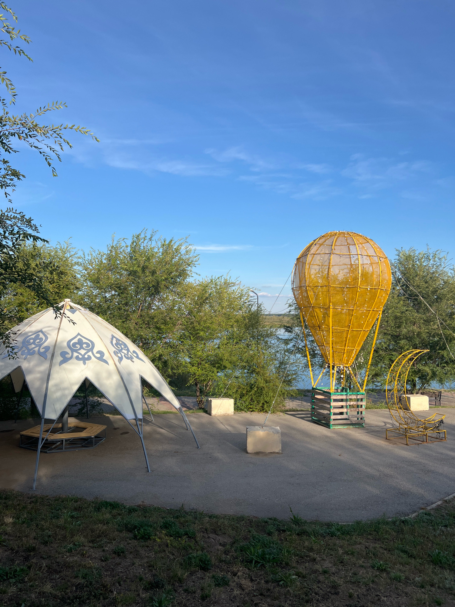 The image depicts an outdoor scene in Kostanay, Kazakhstan, with a clear blue sky and some wispy clouds. In the foreground, there is a unique, white, conical-shaped tent with a geometric pattern on its exterior. Beneath the tent, a hexagonal wooden bench is visible. To the right of the tent, a large, yellow, decorative hot air balloon structure stands on a wooden platform. The balloon is illuminated with small lights and appears to be a permanent fixture. Adjacent to the hot air balloon, a yellow, crescent moon-shaped decoration is also present. In the background, a body of water, likely a river or lake, is visible, bordered by lush green trees. The ground is paved with asphalt, with patches of grass and dry vegetation along the edges. The overall impression is of a park or recreational area. There are no people visible in the image.