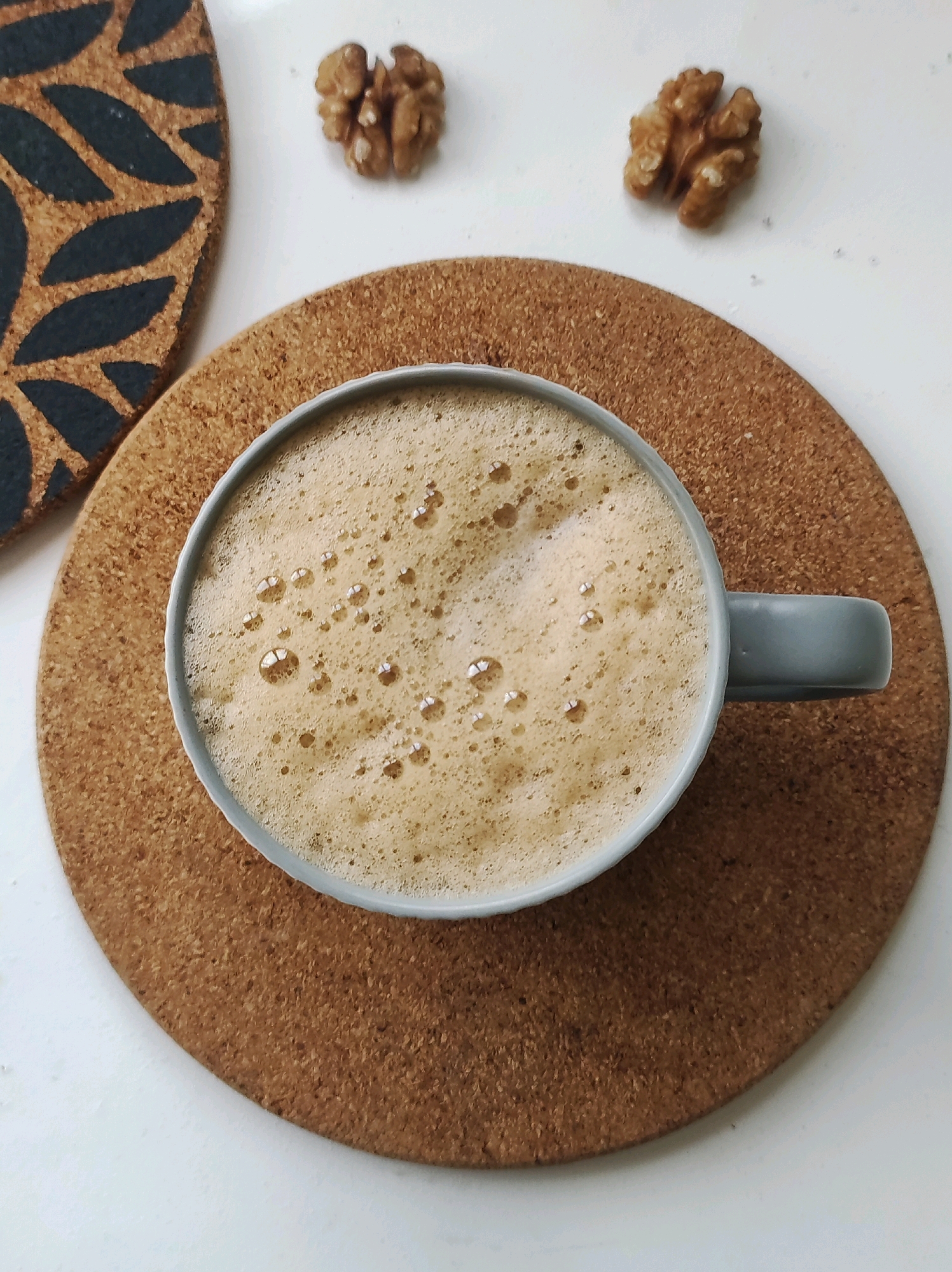 A top-down, medium shot captures a gray mug filled with foamy coffee. The mug rests on a round cork coaster, which is placed on a white surface. To the left of the mug, another cork coaster with a dark leaf pattern is partially visible. Two walnut halves are situated on the white surface above the mug. The scene is brightly lit, casting soft shadows. There are no people present in the image. The image does not provide any information about the city of Chaikovsky, Russia.