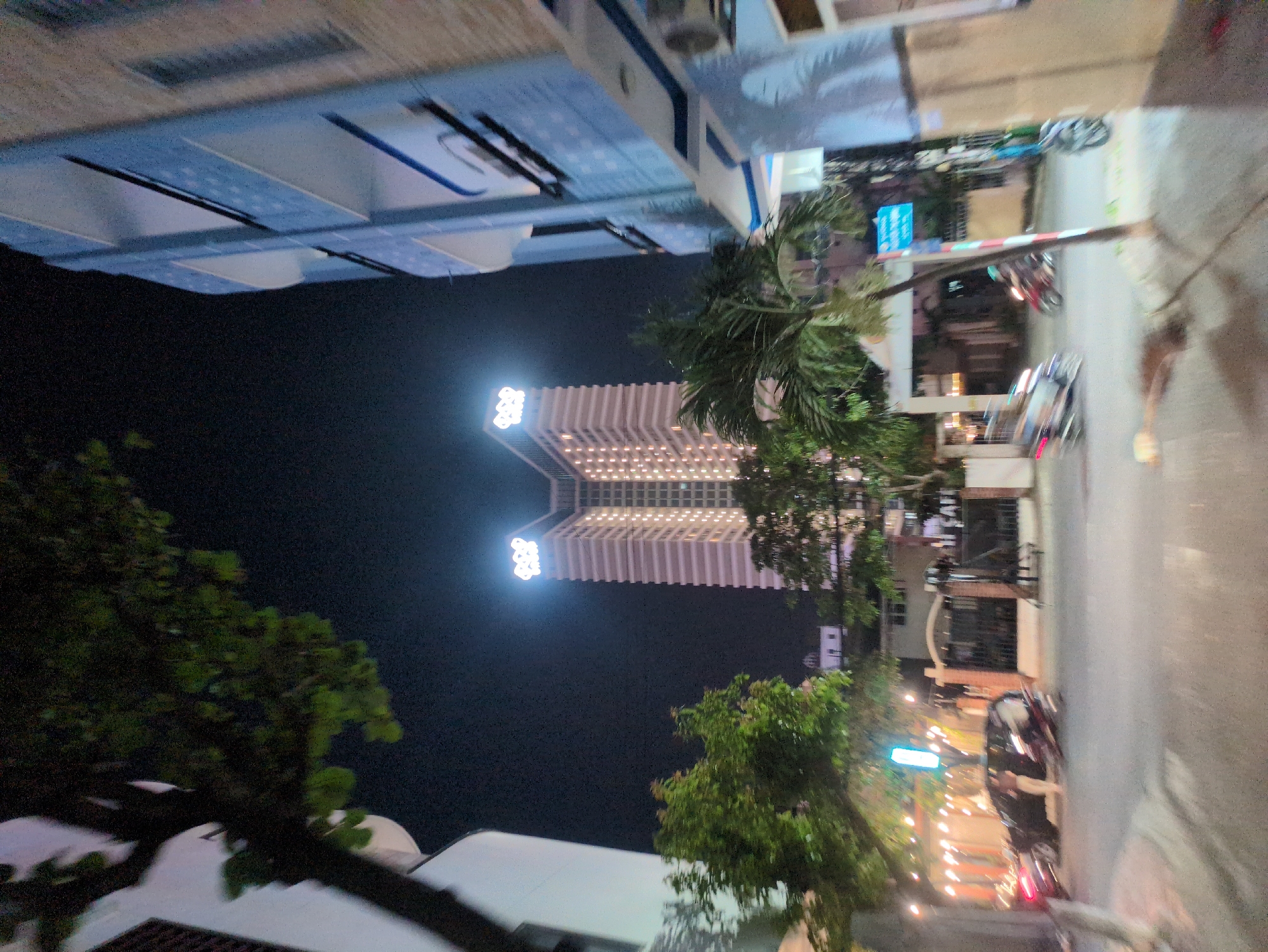 The image depicts a street scene in South Quy Nhon Ward, Vietnam, at night. A tall, brightly lit building is visible in the background, likely a hotel or apartment complex, bearing a distinctive logo at its top. The foreground features trees and the tops of buildings, framing the central view of the illuminated structure. On the right side of the image, a street and sidewalk are present, with glimpses of traffic and streetlights indicating urban activity. The overall impression is of a bustling urban setting with modern architecture.