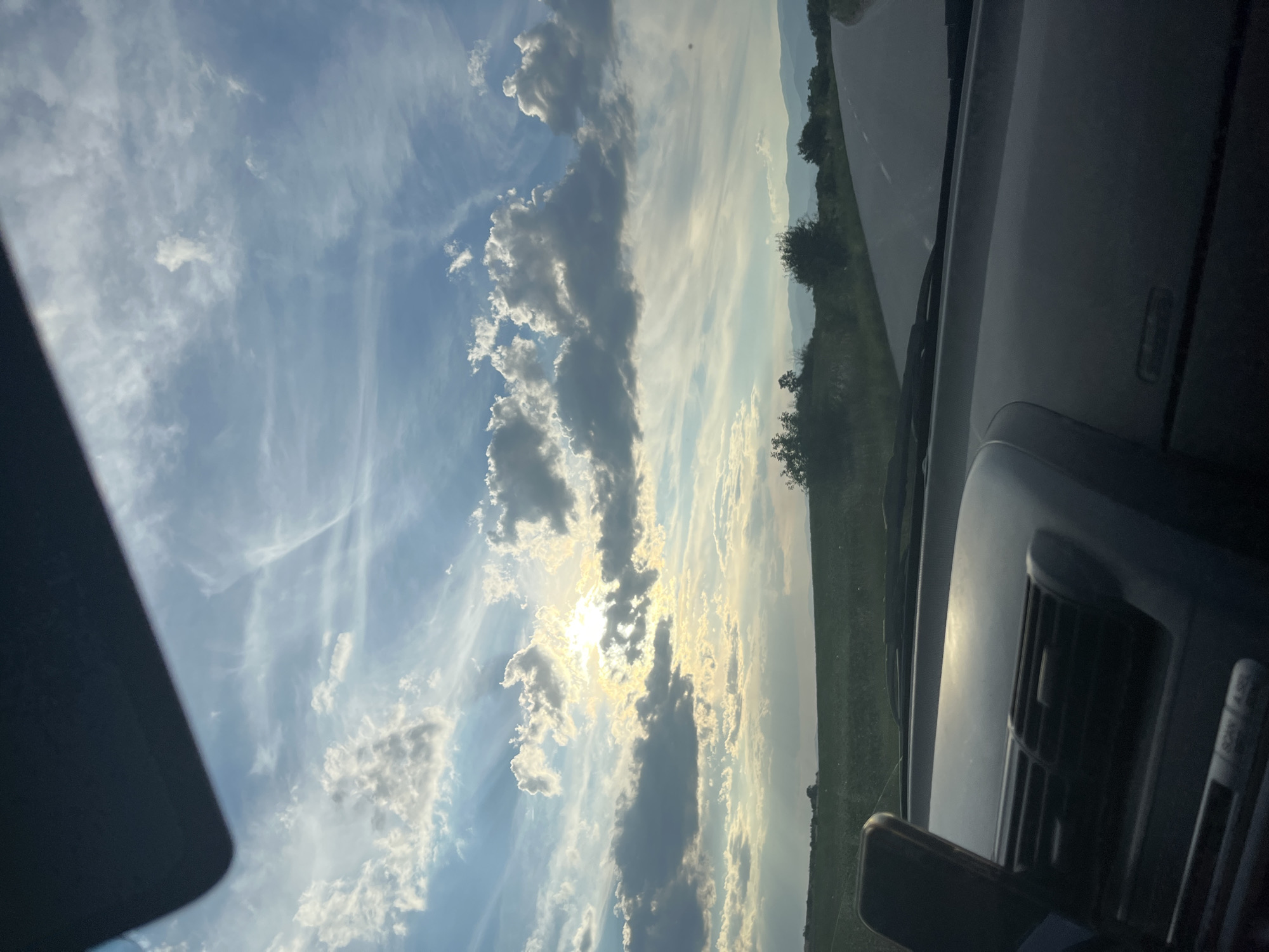 The image captures a view from the interior of a vehicle, looking out at a scenic landscape. The foreground shows the dashboard of the car, with a dark material dominating the lower half of the frame, including what appears to be an air vent and a phone mount. Beyond the vehicle, a paved road curves to the right, leading into a rural setting. The middle ground features a vast, green agricultural field and scattered trees. The background is a vista of rolling hills and mountains under a dramatic sky. The sky is filled with a mix of large, fluffy white clouds and thinner, wispy cirrus clouds, illuminated by the sun which is partially obscured by clouds, creating shafts of light. The overall impression is of a sunny day with dynamic cloud formations, possibly late afternoon or early evening given the light. The scene is consistent with a location in the countryside surrounding Zhitnitsa, Bulgaria, characterized by open fields, hills, and a temperate climate. There are no people visible in the image.