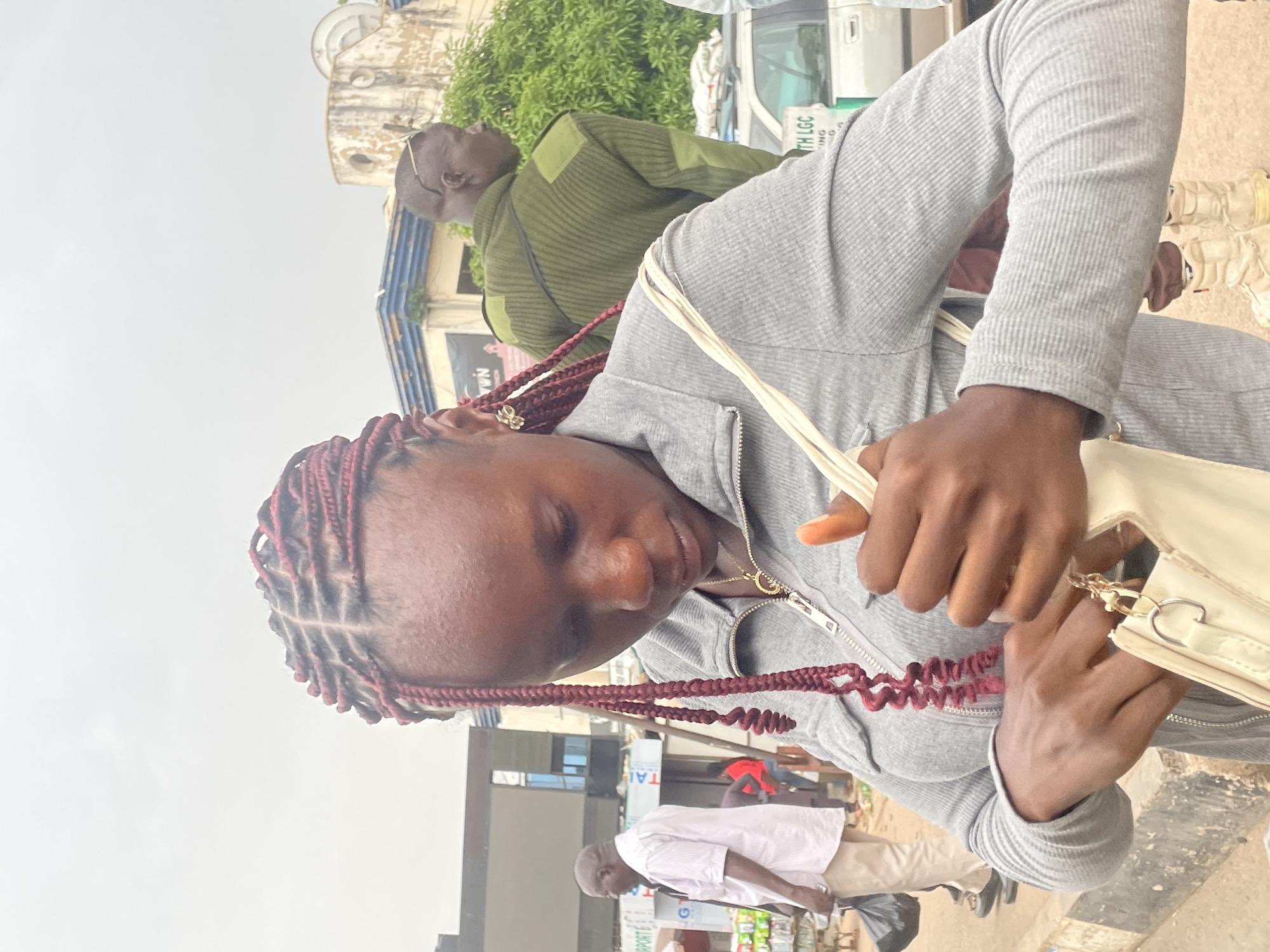 The image shows a woman with red-brown braided hair, wearing a gray ribbed long-sleeved jacket, holding a cream-colored handbag. She appears to be on a street in Jos, Nigeria. In the background, there are people walking, including a man in a green jacket and another man in a white shirt and khaki pants, a building with signage that includes the word "AIRPORT", and a white vehicle with green text "TH LGC". The sky is overcast.