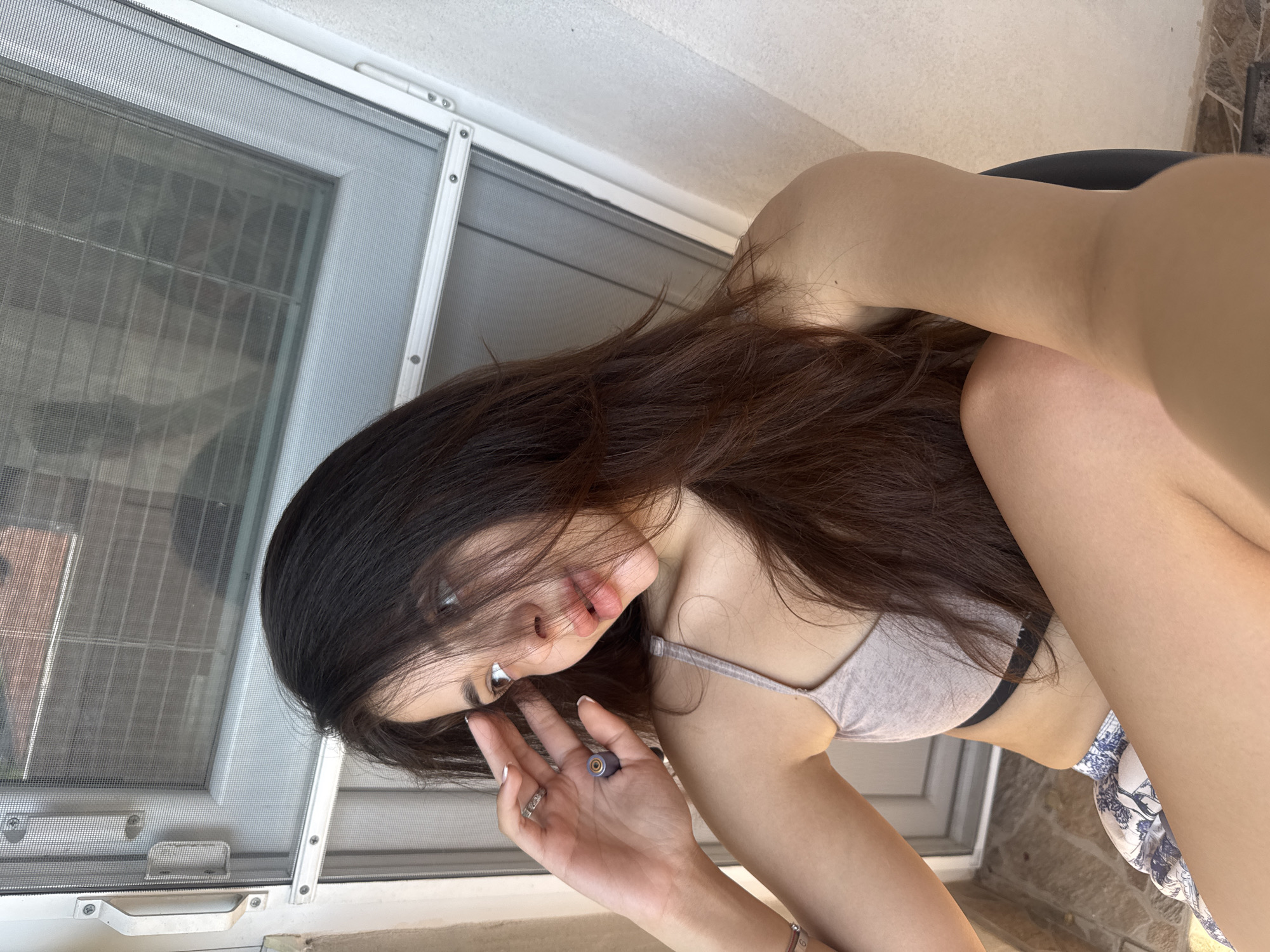 An outdoor selfie shows a young woman with long, dark brown hair sitting on a dark chair near a white-framed window with a screen. The woman is wearing a light-colored bralette and patterned shorts. Her left hand is raised to her forehead, and she is wearing rings on two fingers. The background appears to be a balcony or patio area with tiled flooring and a textured wall. The scene is brightly lit, suggesting daylight. The location is identified as Plovdiv, Bulgaria.