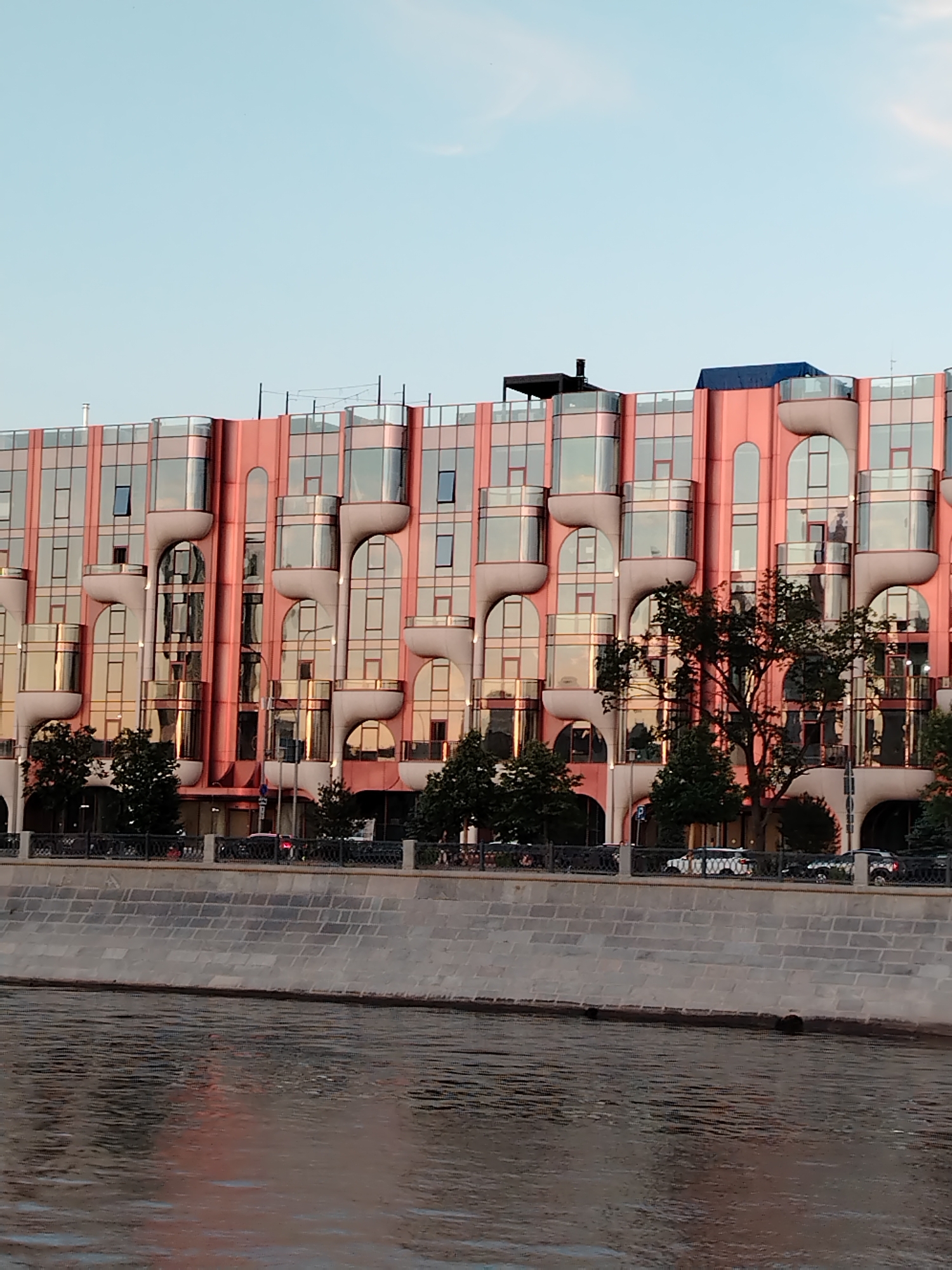 The image shows a modern, pink building with an unusual facade featuring a series of curved, protruding window sections. The building overlooks a wide river. The windows are reflective, showing the sky and surrounding elements. Trees line the embankment in front of the building, and a decorative metal fence separates the walkway from the river. Cars are visible on the road behind the fence, suggesting an urban setting. The sky is clear and blue, indicating daytime, likely late afternoon or early evening given the warm tones in the building's reflection. The water of the river is dark and choppy, reflecting some of the pink hues from the building. The architecture of the building is distinctive, with rounded elements and a layered appearance. The overall scene is one of urban architecture juxtaposed with a natural element, the river. There are no discernible people in the image, and no visible text. The context provided places the scene in Moscow, Russia.