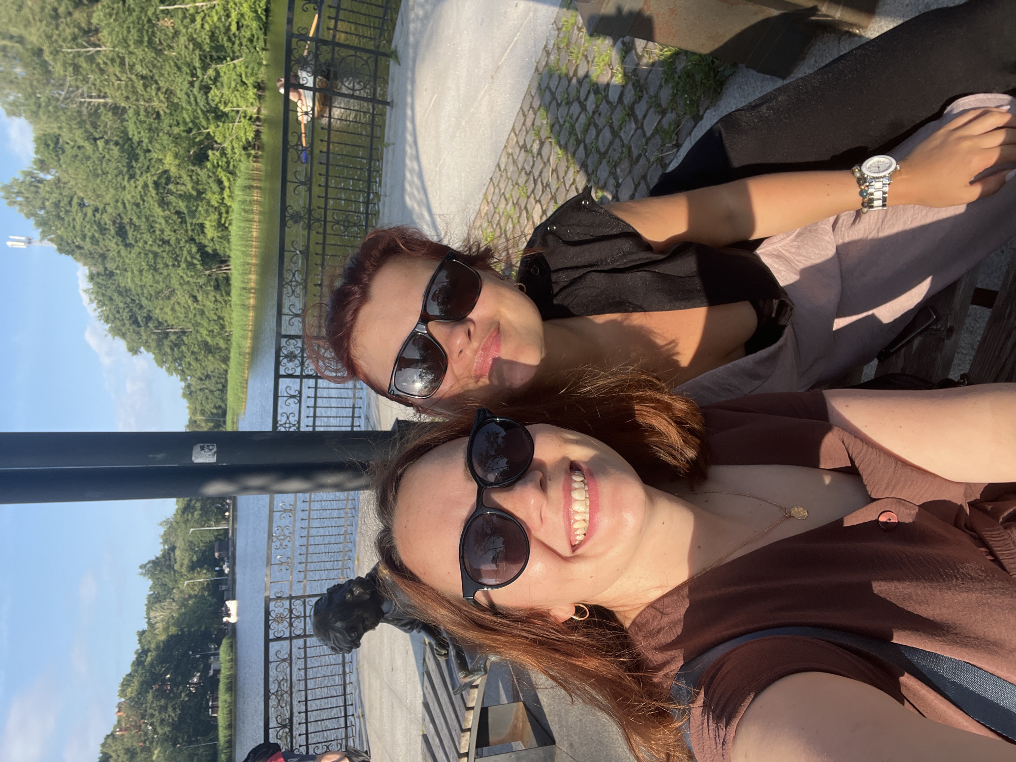 In Svetlogorsk, Russia, two women are seated on a park bench near a body of water, possibly the Baltic Sea. The woman in the foreground, on the left, has long auburn hair and is wearing sunglasses and a brown blouse. She is smiling at the camera and has a gold necklace with a pendant. The woman behind her, on the right, also wears sunglasses and a black top. They are positioned in front of a dark metal railing that borders the water. In the background, lush green trees line the opposite shore, and a few boats, including a swan-shaped paddle boat and a rowing boat with two occupants, are visible on the water. The sunlight casts long shadows, indicating it is either early morning or late afternoon. A small bronze statue of a child is visible to the left of the women, also seated on a bench. The ground is paved with cobblestones, and there is some greenery growing between the stones.