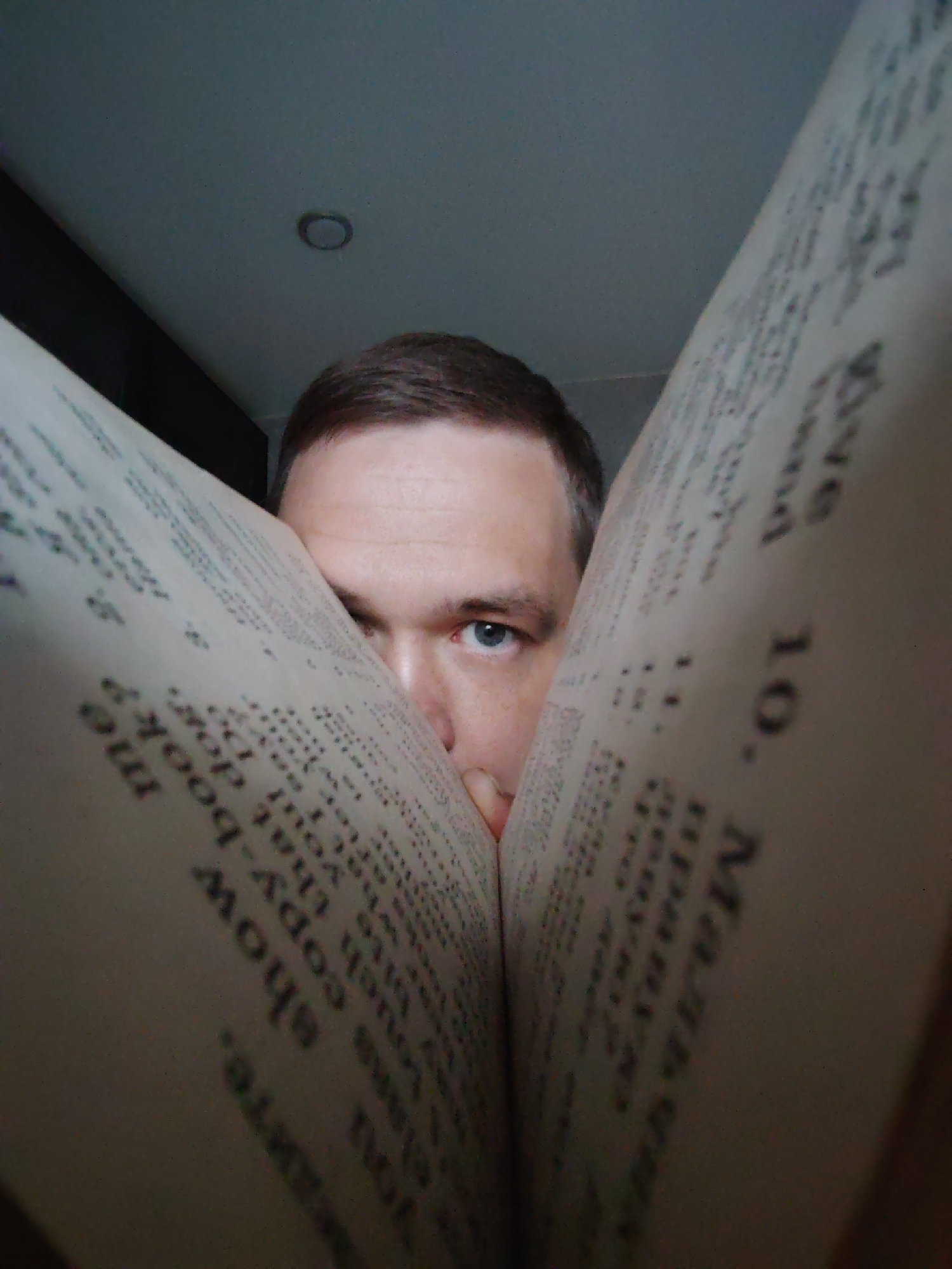 A close-up image shows a person with fair skin and short brown hair holding a book open in front of their face. Only their right eye and part of their nose are visible between the pages. The book's pages are filled with text in a dark font, including the words "you show me" and "copy-book?" and "10. Мальчик". The background is a neutral-colored ceiling with a recessed light fixture. There is no indication of Chaikovsky, Russia in this image.