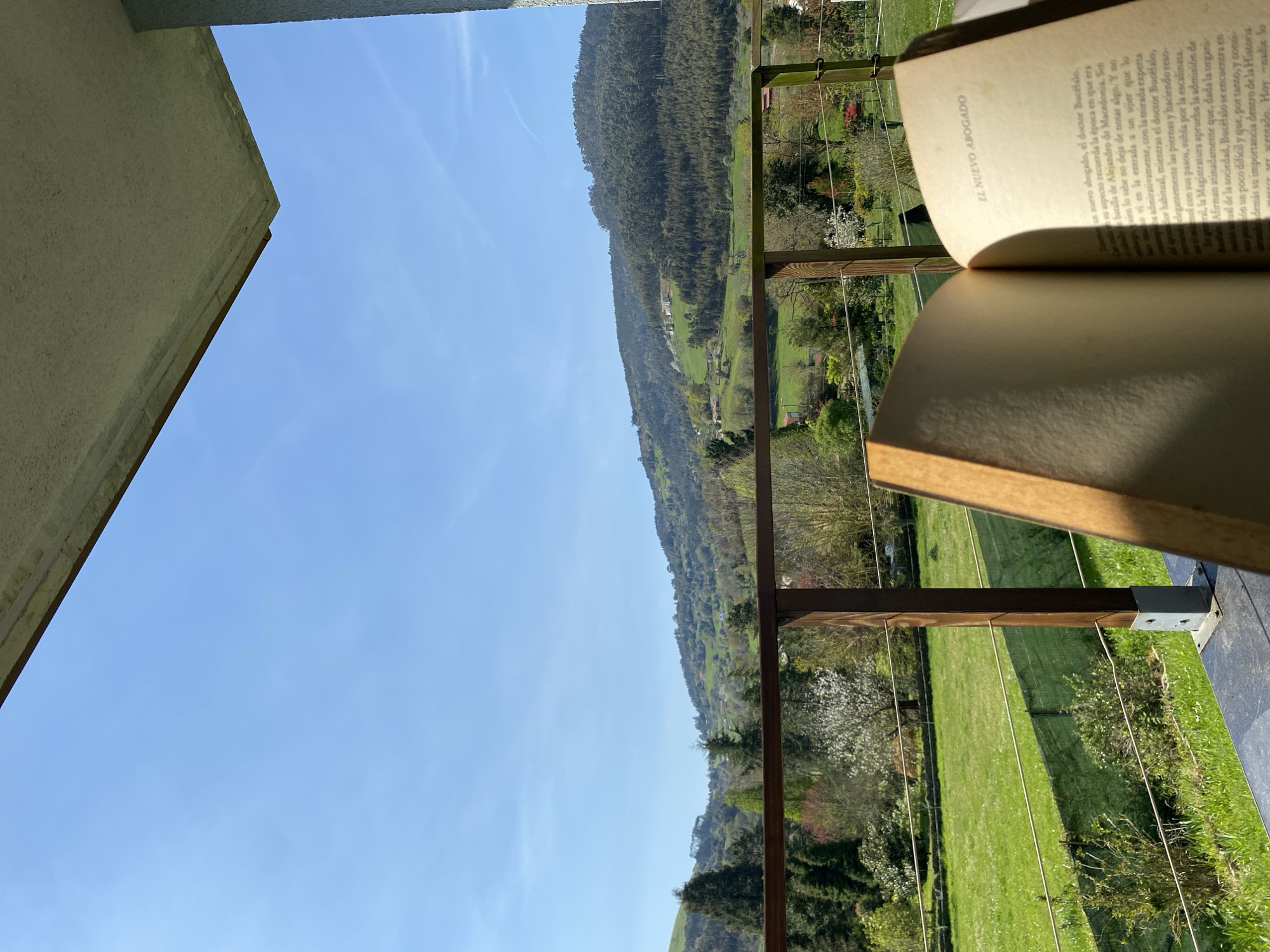 The photograph depicts an open book titled "El Nuevo Abogado" resting on a balcony, possibly in a residential setting. In the background is a landscape with green fields and a tree-covered mountain. The clear blue sky with faint clouds, the style of the building, and the vegetation suggest the image was taken in a rural area. The geographical location is specifically Villaviciosa, Spain.