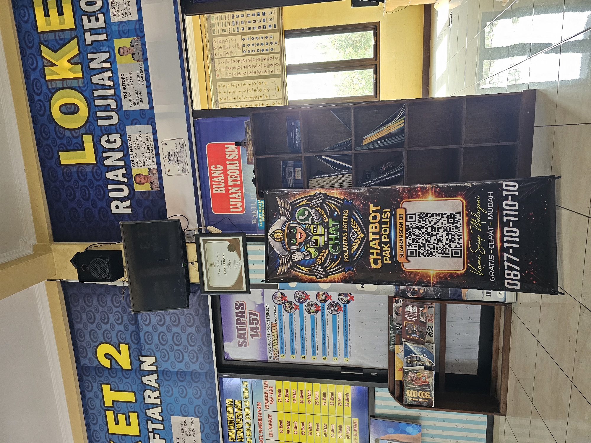 The image depicts the interior of what appears to be a government office or a service center, possibly related to driver's license applications, given the text "LOKER RUANG UJIAN TEORI SIM" (Job Vacancy Theory Exam Room SIM) visible on a banner. The location is identified as Madureso, Indonesia through the banner text "POLRES TERANGGUNG".
Dominating the scene is a large black banner featuring a cartoon police officer with the text "CHAT POLANTAS JATENG" and a QR code, along with a phone number "0877-110-110-10" and the slogan "Kami Siap Melayani GRATIS - CEPAT - MUDAH". To the left of this banner is another smaller banner displaying a table with time durations like "60 Menit", "90 Menit", and "25 Menit", suggesting service times.
Further to the left, a large blue banner with yellow text reads "KET 2" and "FTARAN". Mounted on the wall above this banner is a black flat-screen television. Above the television, a black speaker is mounted on the wall.
A wooden bookshelf filled with books and magazines is visible to the right of the central banner. Behind the bookshelf is a window with wooden frames, offering a view of the outdoors, with sunlight casting reflections on the tiled floor.
Several framed documents and certificates are displayed on the wall, including one with text in Indonesian and what appears to be a police seal. Two smaller framed portraits of men in uniform are also visible on the wall, part of the "RUANG UJIAN TEORI SIM" banner, with names and ranks listed.
The overall environment appears to be a public service area with informational displays and amenities for visitors. The lighting suggests it is daytime, with natural light entering through the window. The floor is tiled in a light color.