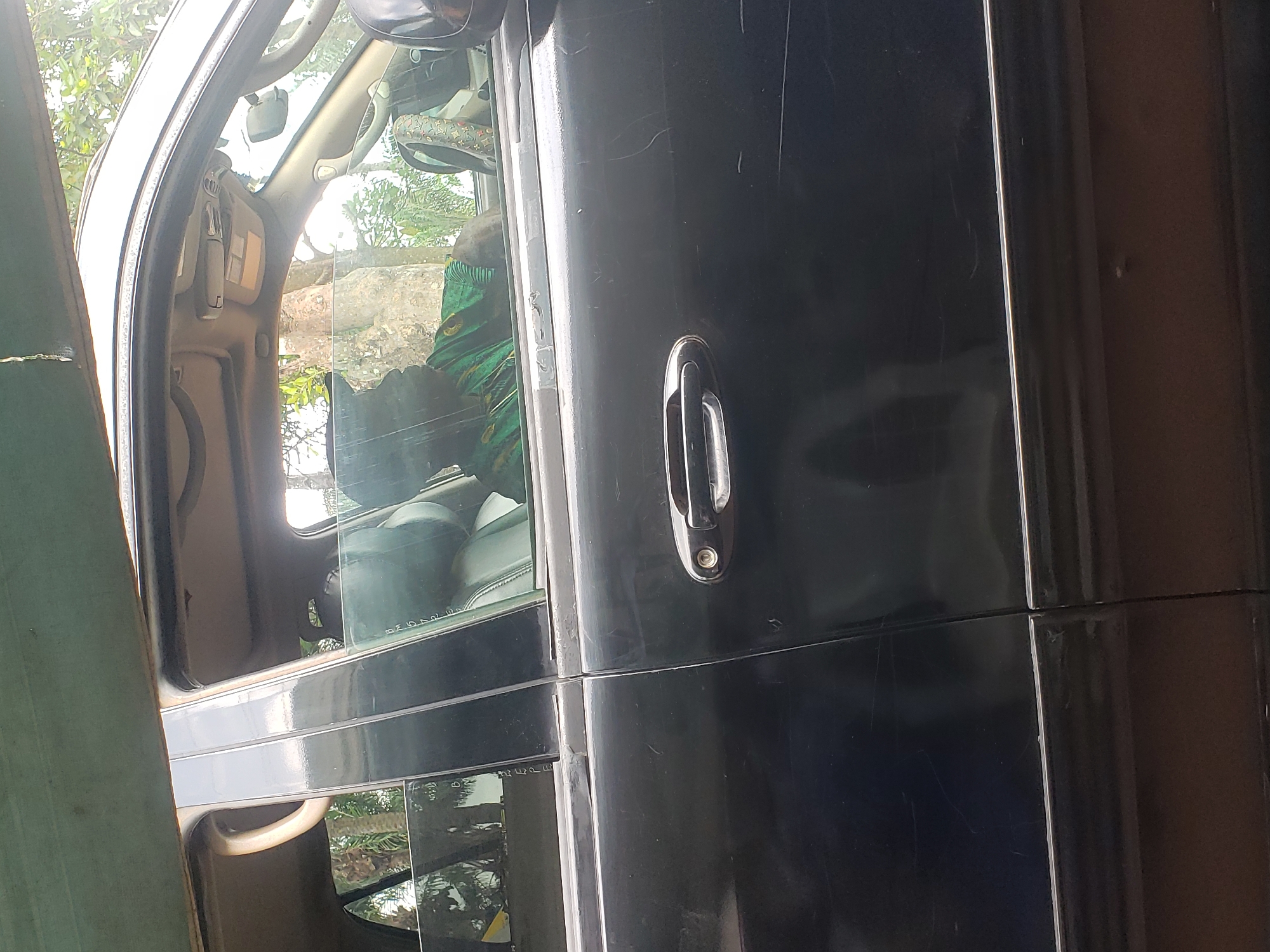 The image shows the side of a black vehicle, possibly a car or truck, with an open door. The door handle is visible. Through the window, part of the interior of the car is visible, including seats and the steering wheel. The interior appears cluttered with a green and yellow patterned fabric draped over the steering wheel. The background shows a blur of trees and vegetation. The photo was taken in the city of Umundugba, Nigeria.