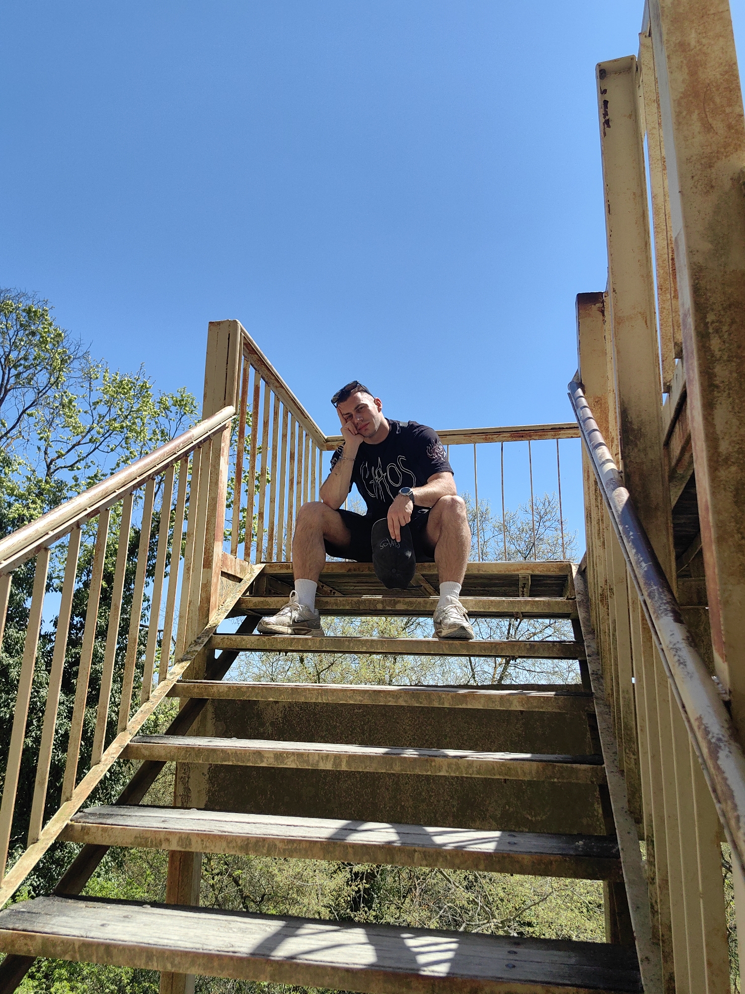 A man sits on rusted metal stairs under a clear blue sky. He wears a dark t-shirt, black shorts, white socks, and light-colored sneakers. He has sunglasses on top of his head and holds a black baseball cap in his hand. Green trees and foliage are visible in the background. The location is likely in Varna, Bulgaria, based on image request.