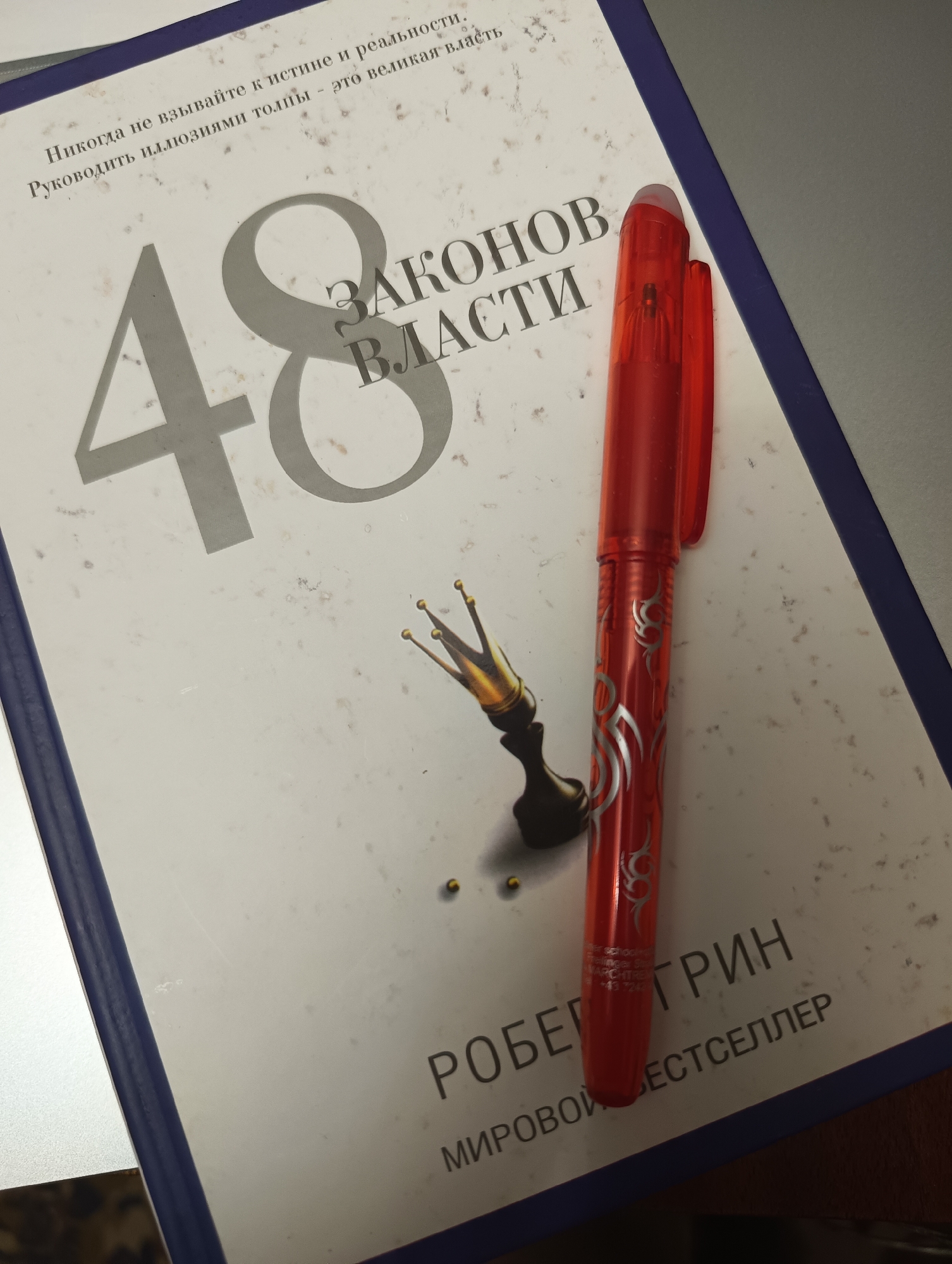 A red pen rests on a book titled "48 Laws of Power" by Robert Greene. The book is open, and the pen is positioned diagonally across the pages. The cover of the book appears to be white with large gray numbering and text. There is an illustration of a golden crown with three prongs on the left side of the book, near the center. The background is a muted, textured surface, possibly a table or desk. The lighting suggests an indoor environment, possibly a home or office. No specific location within a city is discernible.