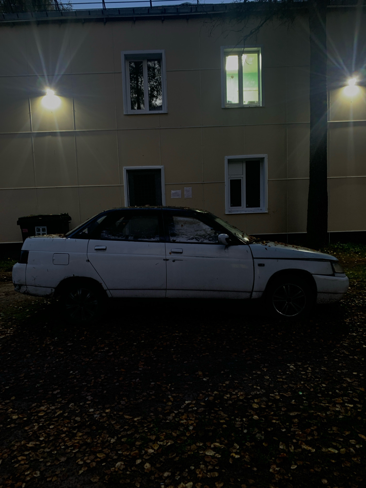 A white sedan, possibly a VAZ-2110, is parked on a dark, leaf-strewn surface in front of a light yellow building. The building has several windows, some of which are lit from within. Outdoor lights on the building cast bright beams. A black trash bin is visible to the left of the car. The scene appears to be in the city of Katunino, Russia.