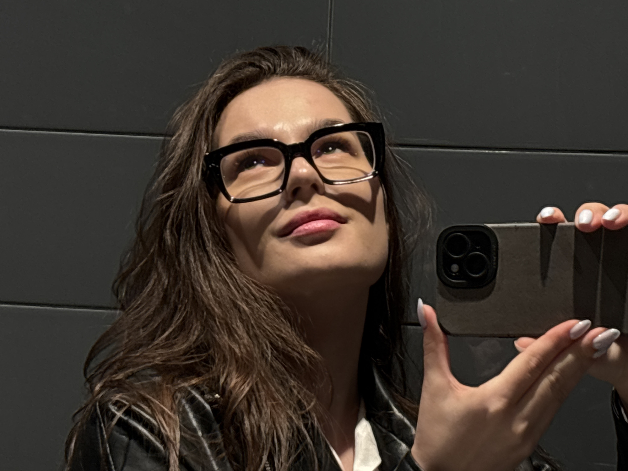 A young woman is captured in a self-portrait, holding a smartphone in her right hand as she gazes upwards. She has long, dark brown hair that falls around her shoulders and is wearing large, black-framed glasses. Her lips are slightly parted, and a soft shadow from her glasses is cast across the upper part of her face. She appears to be wearing a dark, possibly leather, jacket over a white collared shirt. Her fingernails are manicured with a light, white polish in an almond shape.
The smartphone she holds has a dark-colored case, and the triple camera lens array is clearly visible on the back. The background is a plain, dark gray wall with a horizontal seam, suggesting an indoor setting. The overall lighting is soft, highlighting her features and creating a gentle reflection on her glasses. The activity is clearly a selfie, with the woman looking into the phone's lens or screen held just out of frame. There is no visible text in the image. The context provided indicates the location is Izhevsk, Russia.