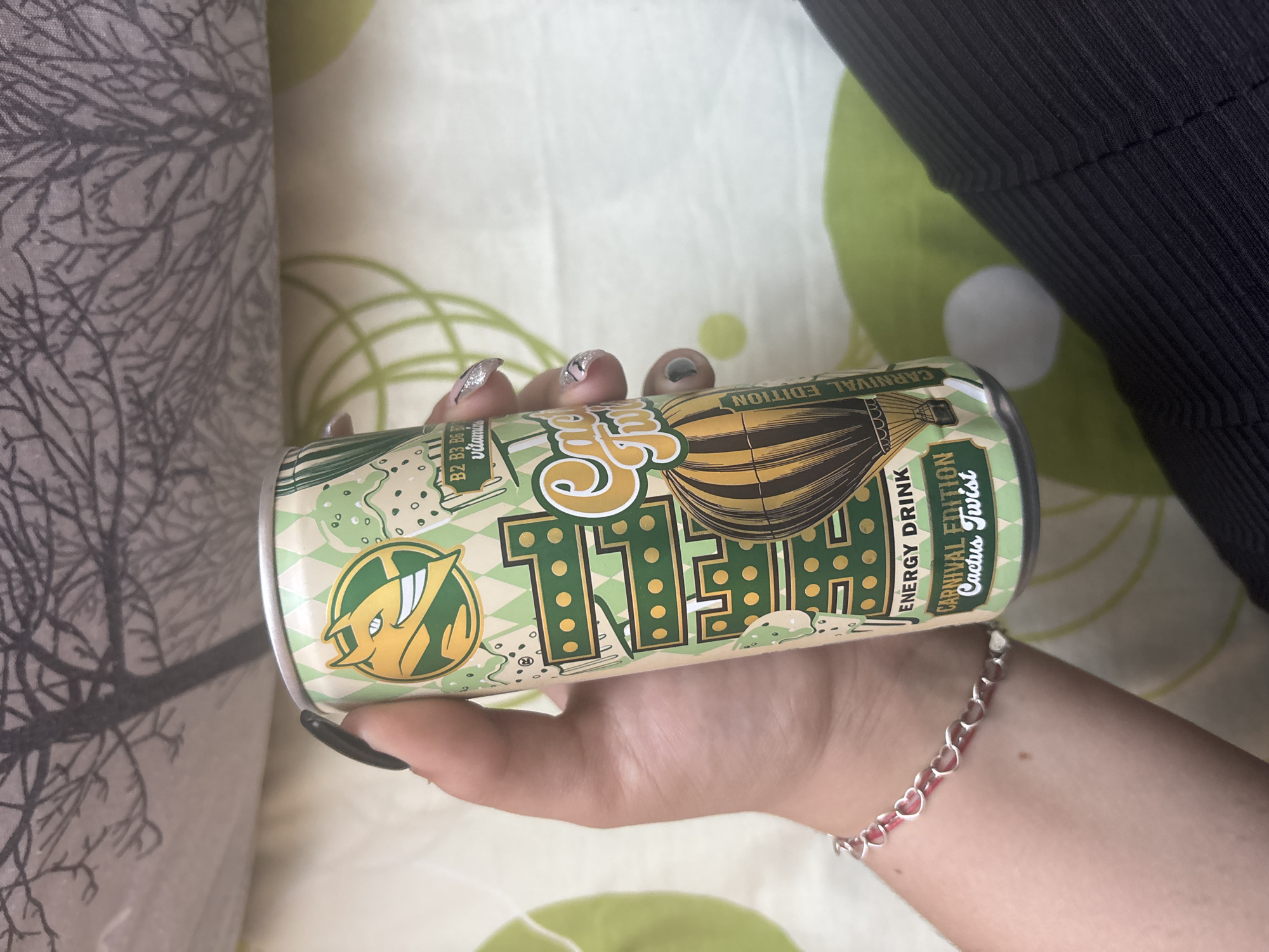 The image shows a person's hand holding a can of "11 TEAM ENERGY DRINK CARNIVAL EDITION Cactus Twist". The can is predominantly green and yellow with an illustration of a hot air balloon and a cartoonish character. Text on the can includes "11 TEAM", "ENERGY DRINK", "CARNIVAL EDITION", and "Cactus Twist". The background consists of a patterned fabric, possibly a blanket or bedding, with abstract tree-like designs and circular green shapes. A dark ribbed fabric, likely clothing, is visible in the upper right corner. The person is wearing a silver bracelet with small red elements and heart shapes. The overall lighting suggests an indoor setting. The photo was taken in Lom, Bulgaria.