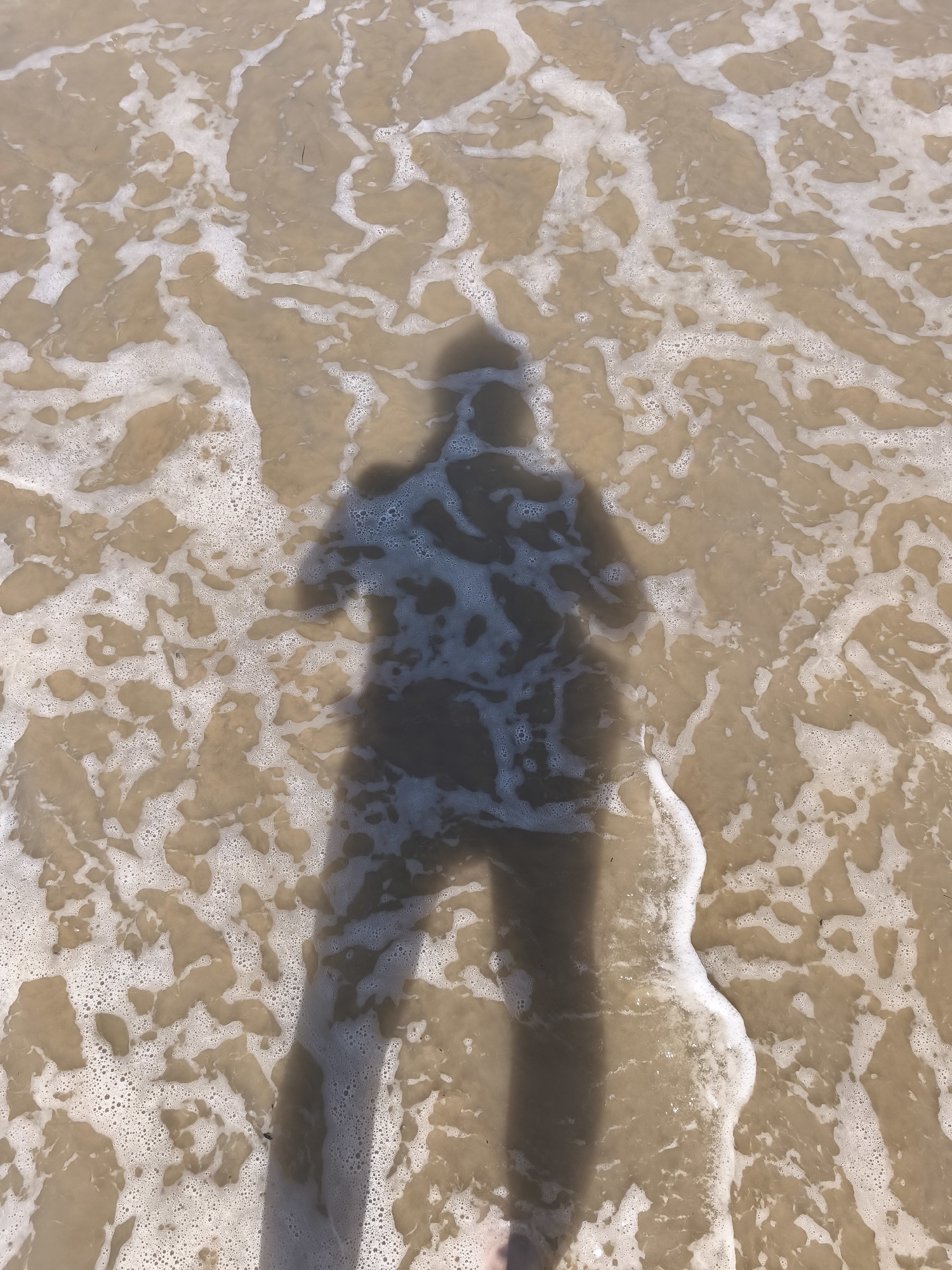An overhead view captures the shadow of a person standing on a sandy beach in San Salvo, Italy, as a gentle wave washes over their feet and the sand. The water is shallow and translucent, revealing the sandy seabed beneath. The wave creates a frothy, foamy edge as it recedes, leaving behind intricate patterns of bubbles and wet sand. The overall impression is one of a tranquil moment on a sunny day at the coast, with the shadow serving as a solitary human presence against the natural elements.
