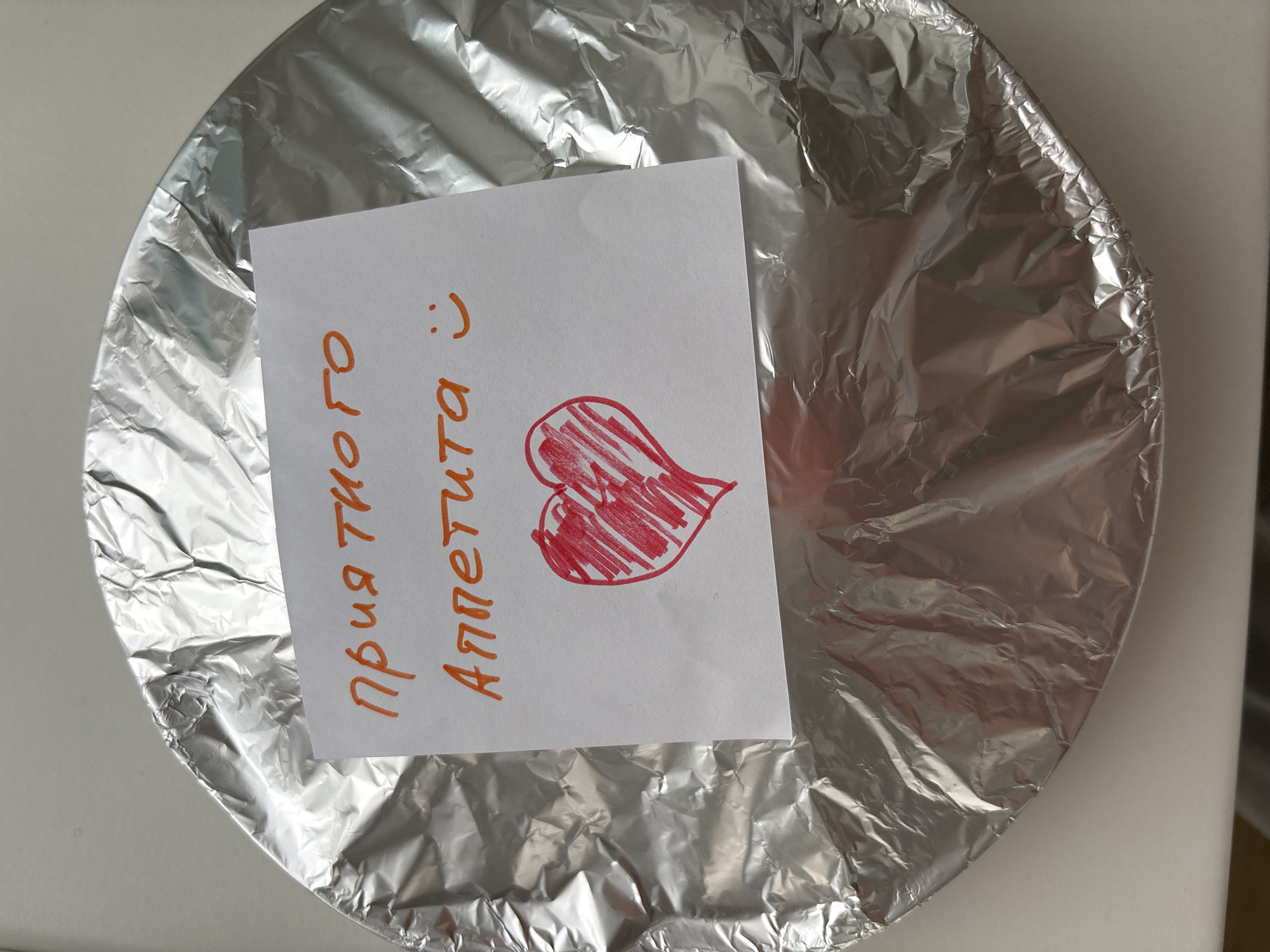 A round object, likely a dish or container, is prominently featured, covered entirely with shiny, crinkled aluminum foil. The foil shows numerous folds and reflections, suggesting it's tightly wrapped around the item. Centered on top of this foil-covered object is a rectangular piece of white paper, serving as a handwritten note.
The note contains text and a drawing, written in what appears to be an orange or red marker. The text reads "приятного Аппетита :)" which translates from Russian to "Enjoy your meal :)" or "Bon appétit :)". Below the text, a hand-drawn heart is visible, outlined in red and filled with red scribbles. The smiley face and the heart convey a warm and friendly sentiment.
The object rests on a light-colored surface, possibly a table or counter, against a subtle background wall. The lighting suggests an indoor setting, likely during the daytime. The overall scene depicts a prepared food item, thoughtfully presented with a personal message, ready to be enjoyed. The location provided for context is Chaikovsky, Russia.
